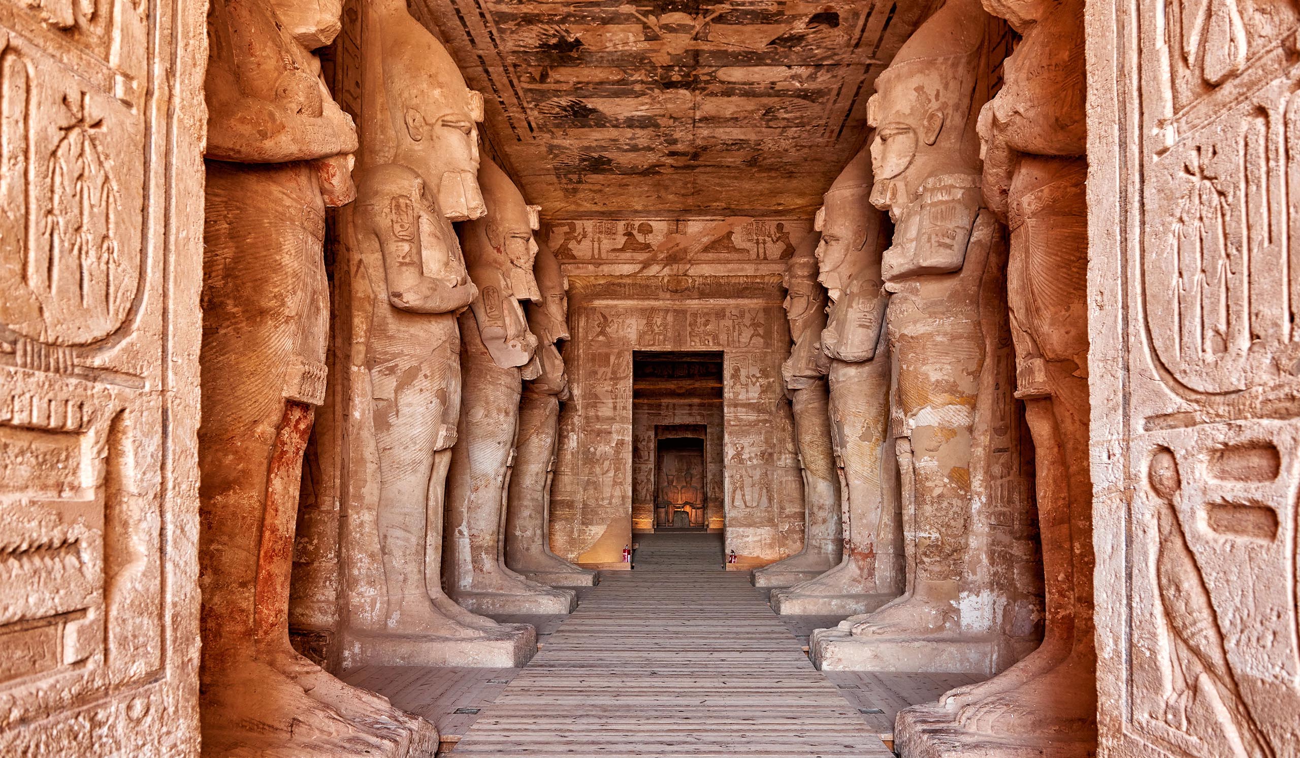 Interior corridor of an ancient Egyptian temple lined with large carved statues and hieroglyph-covered walls, leading to a doorway in the distance.