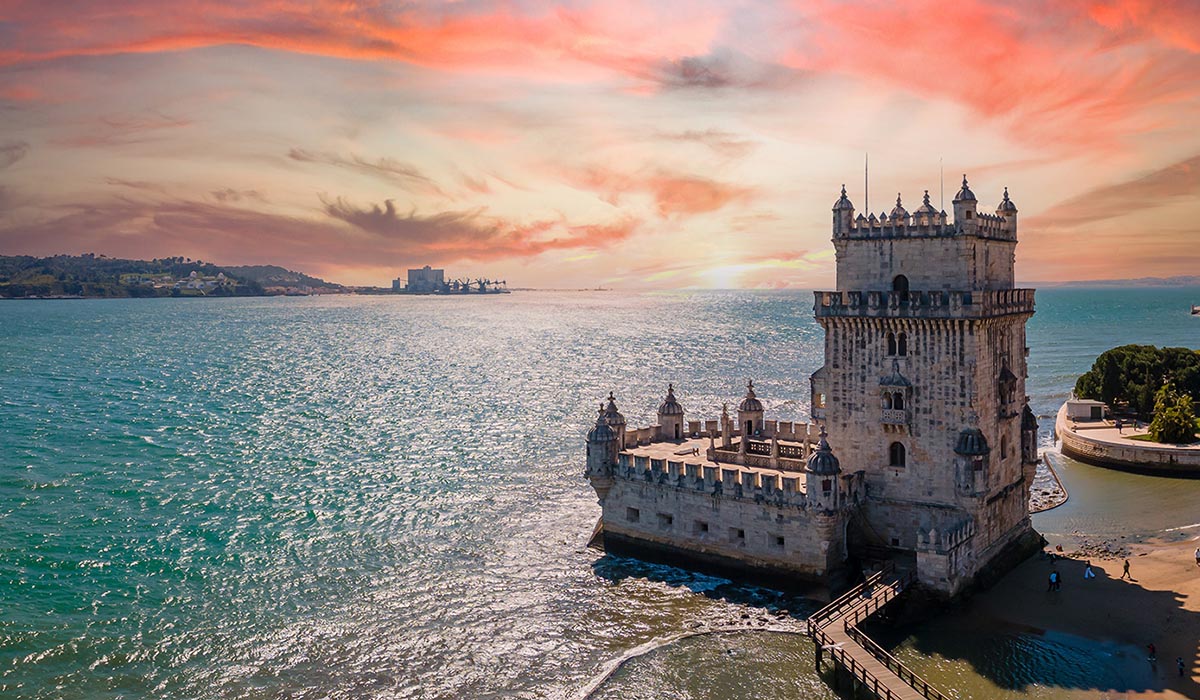 Historic stone tower fortress extending into the water at sunset, with a walkway leading to it and shimmering ocean under a colorful sky.