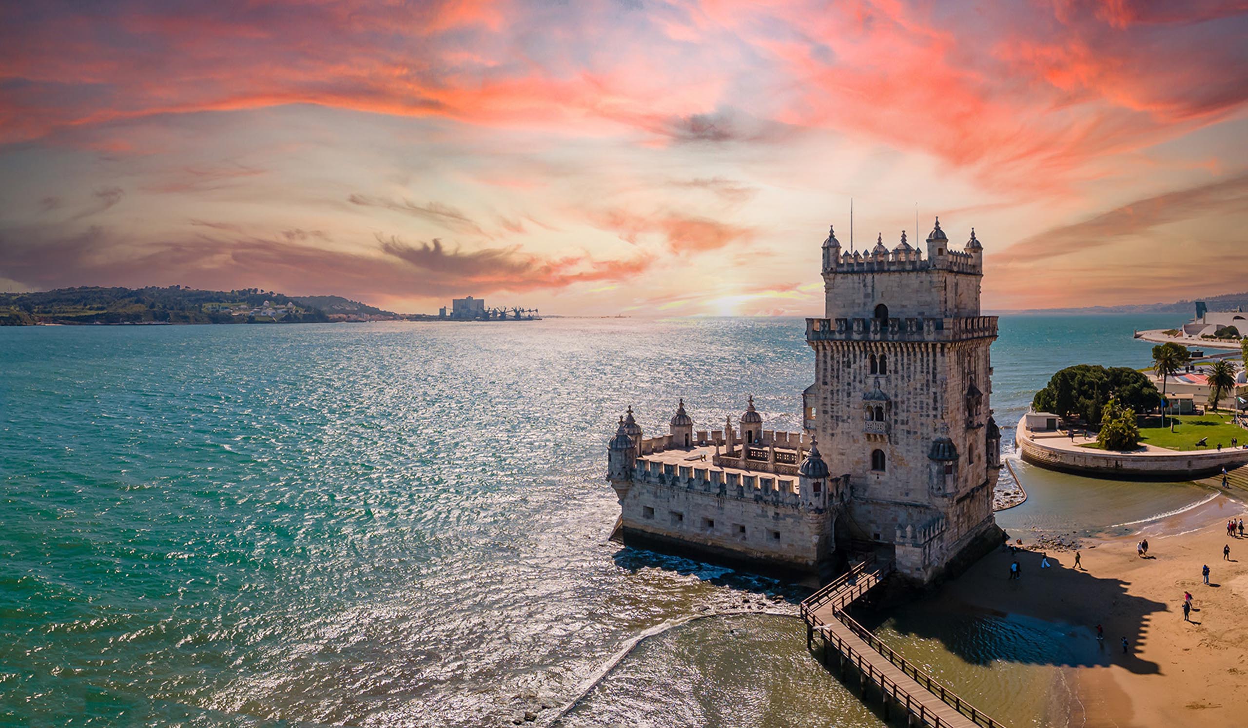 Historic stone tower fortress extending into the water at sunset, with a walkway leading to it and shimmering ocean under a colorful sky.