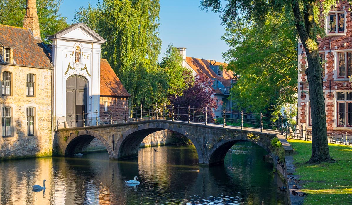 Stone bridge arches over a calm canal beside historic buildings and leafy trees, with reflections in the water.