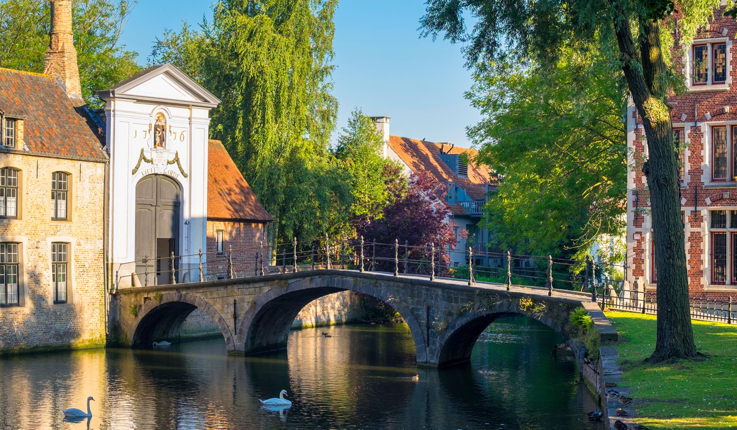 Stone bridge arches over a calm canal beside historic buildings and leafy trees, with reflections in the water.