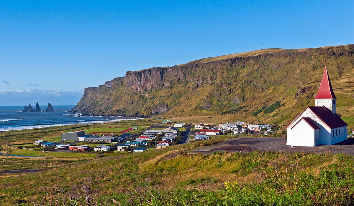 Small coastal village sits beneath steep cliffs by the ocean, with a white church with a red roof in the foreground.