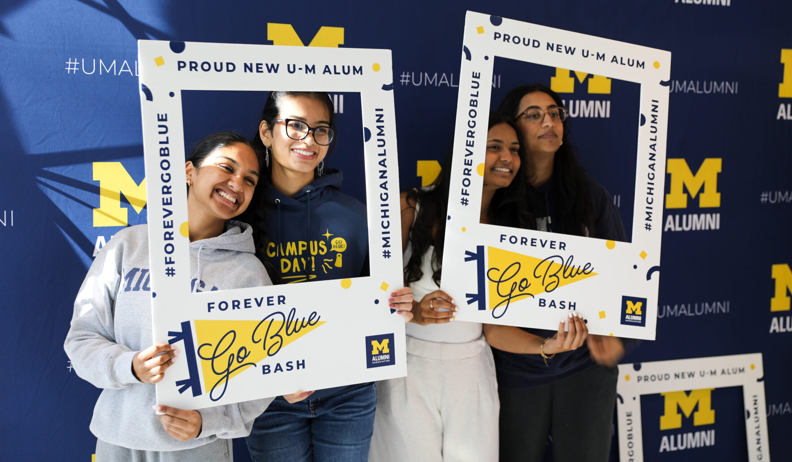 Four smiling people pose together, holding large photo frames that say Forever Go Blue Bash at a University of Michigan End of Semester alumni event. The background features repeated Michigan alumni logos and hashtags.