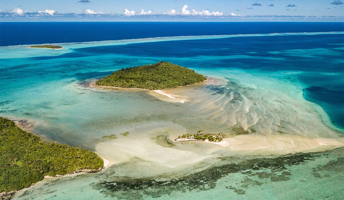 Aerial view of a small tropical island surrounded by turquoise water, with sandbars and shallow reef patterns visible in the lagoon.