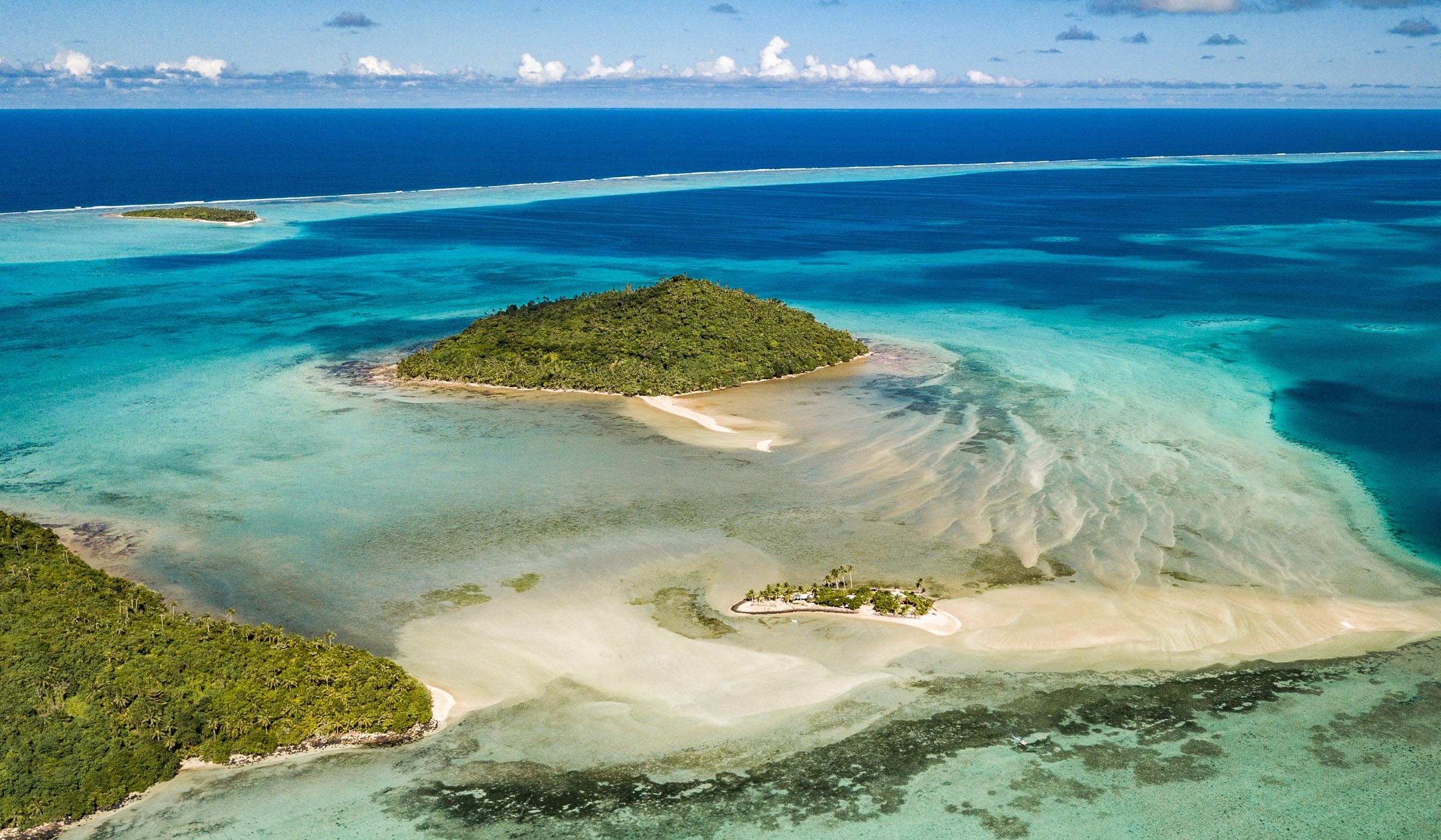 Aerial view of a small tropical island surrounded by turquoise water, with sandbars and shallow reef patterns visible in the lagoon.