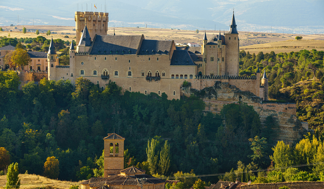 Large stone castle with towers and pointed spires sits atop a wooded cliff, overlooking a valley with open fields in the distance.