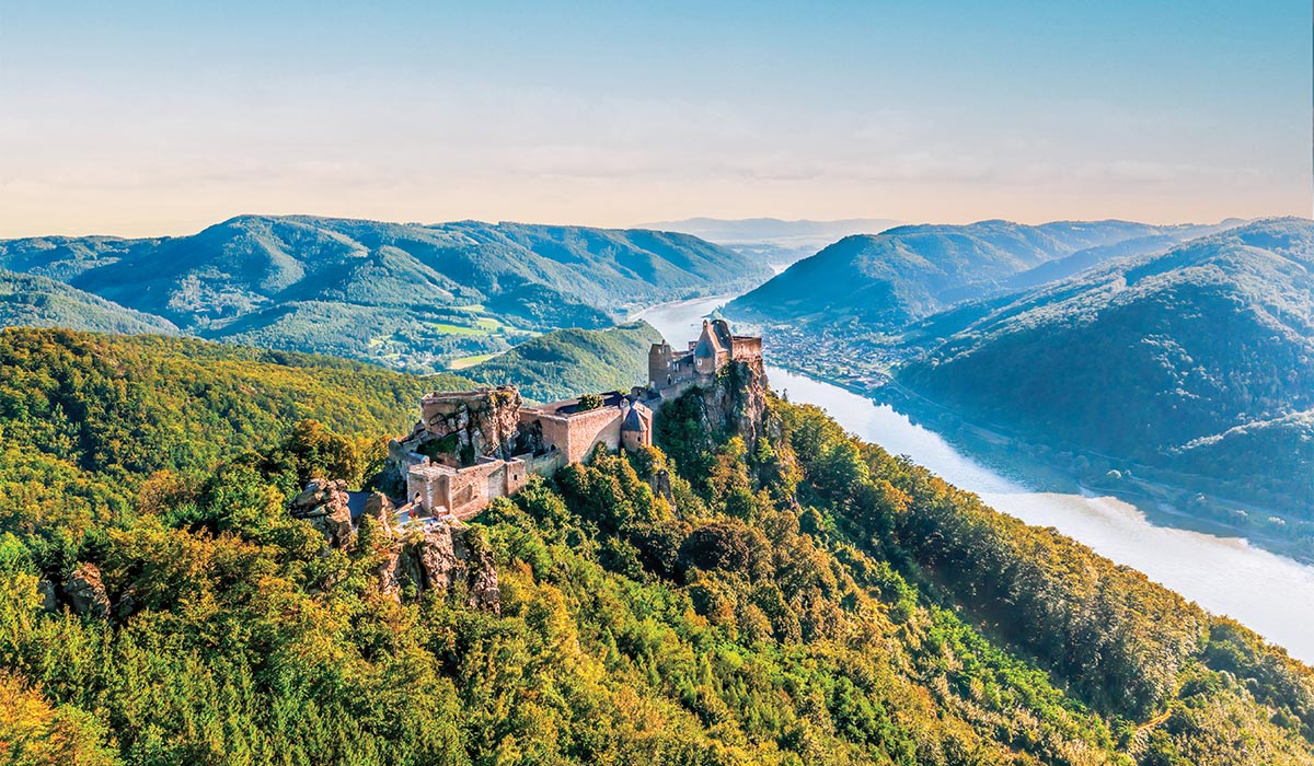 A hilltop castle overlooks a river winding through green mountains under a clear sky.