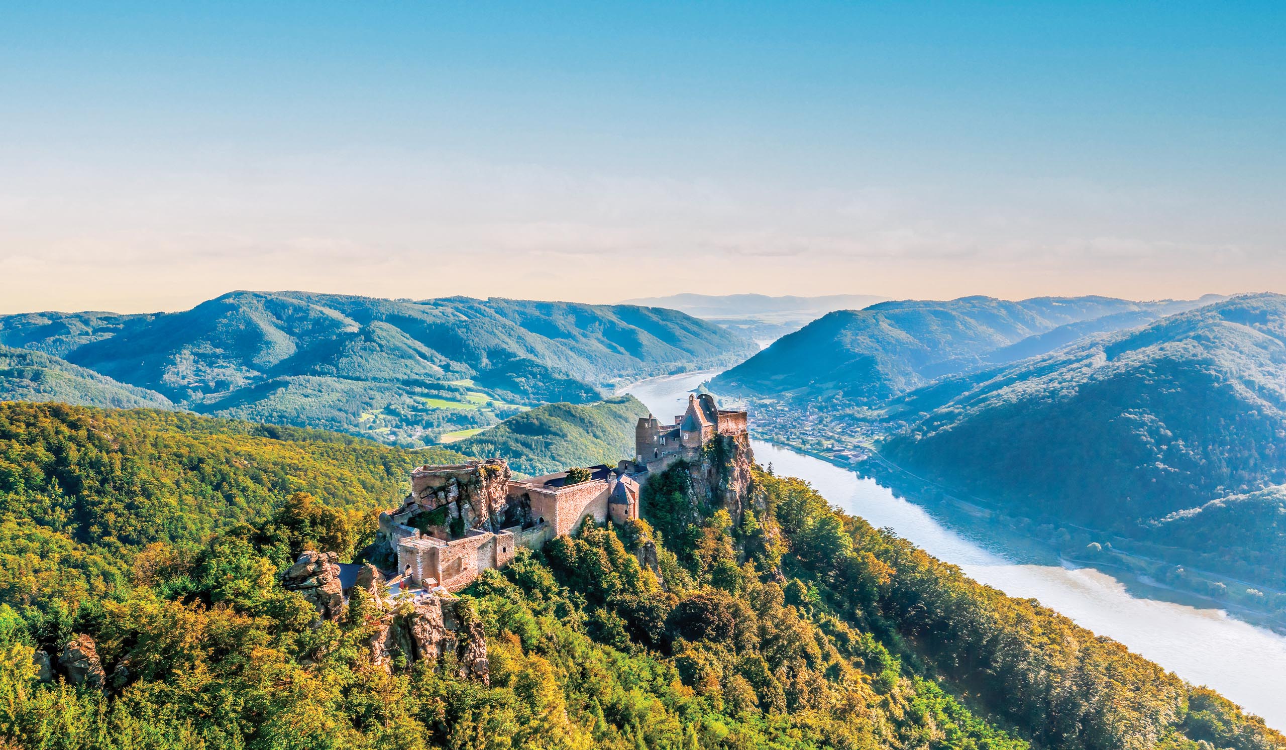 A hilltop castle overlooks a river winding through green mountains under a clear sky.