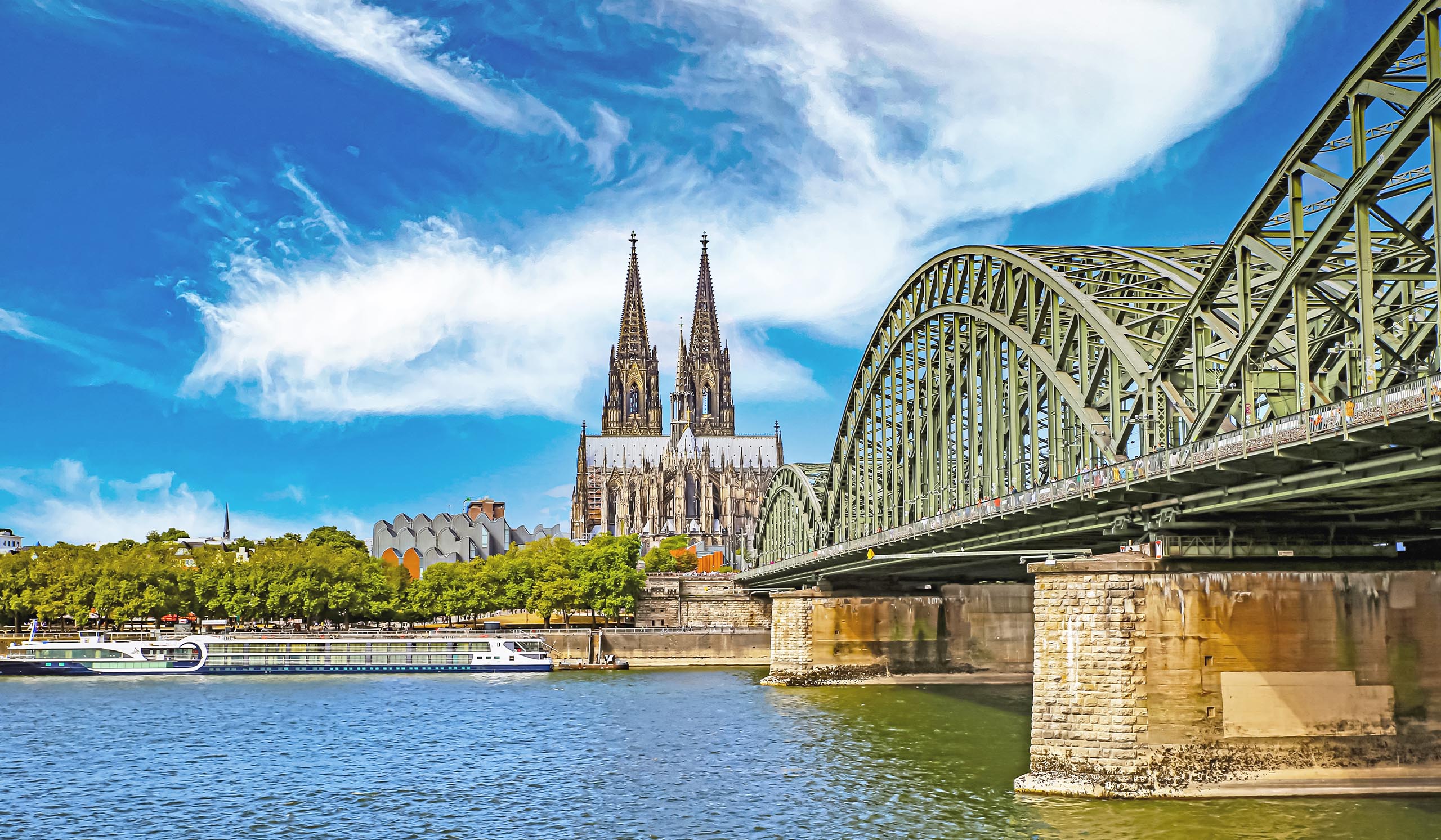 View of a large Gothic cathedral with twin spires behind a steel arch bridge spanning a river, with a boat passing along the water under a bright blue sky.
