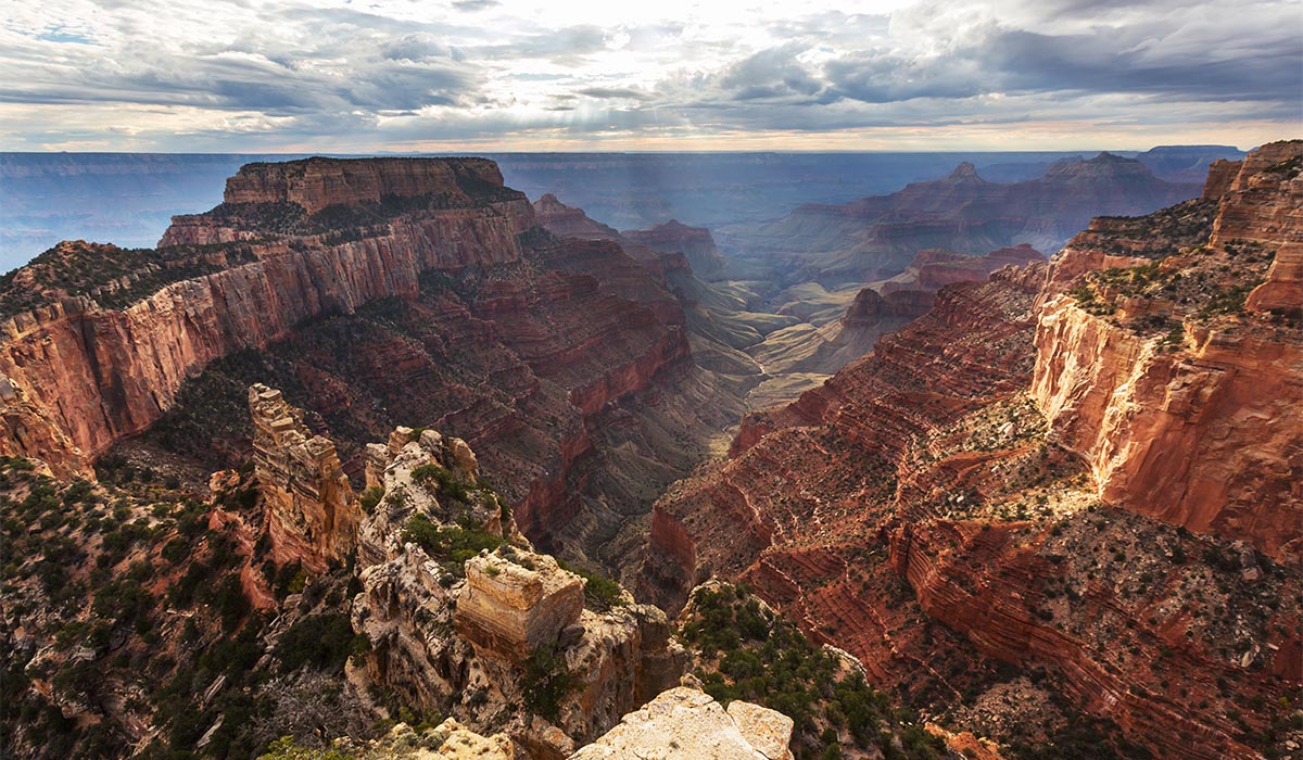 Wide view of the Grand Canyon, with layered red rock cliffs and a deep valley stretching into the distance beneath a cloudy sky with rays of sunlight breaking through.