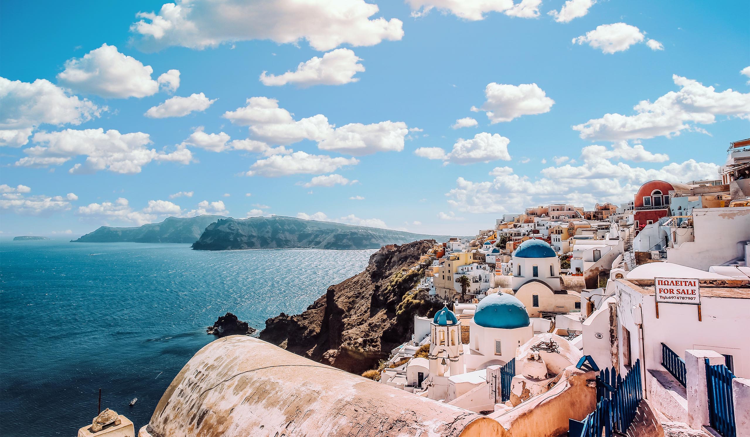 White buildings with blue domes sit on a cliff overlooking the ocean, with a mountainous island in the distance under a bright sky.