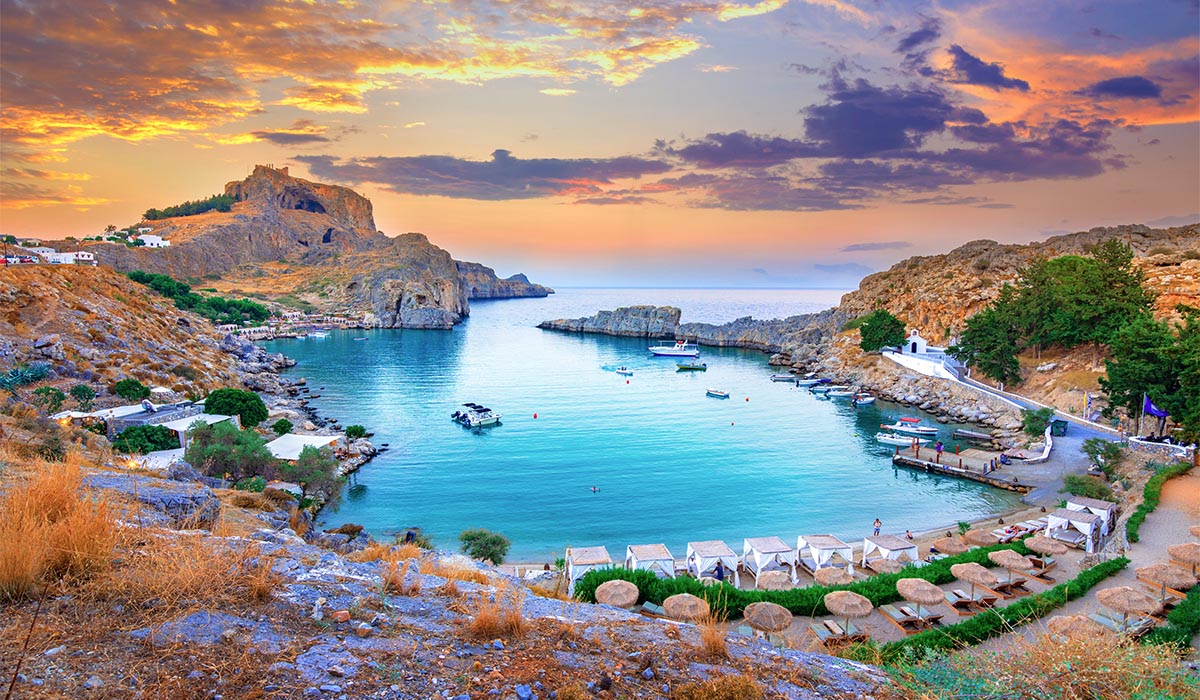 A small bay with clear blue water is surrounded by rocky hills, with boats and a beach below a colorful sunset sky.