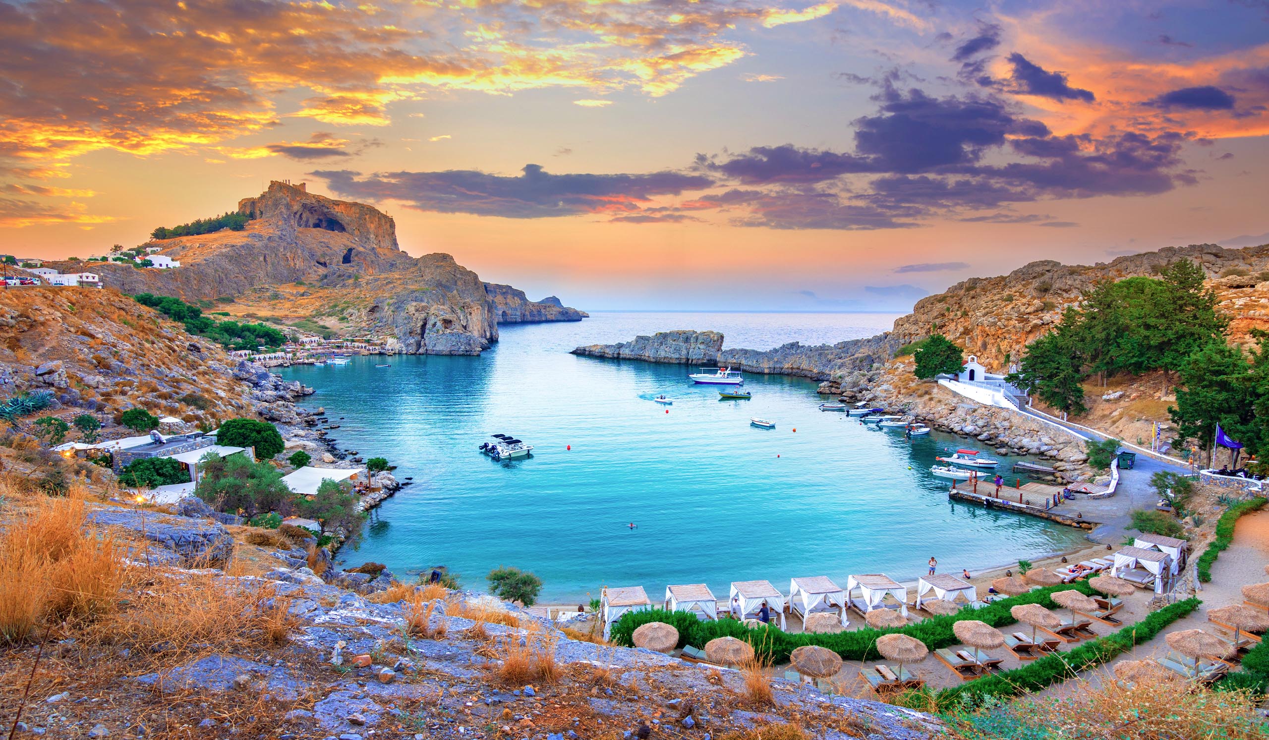 A small bay with clear blue water is surrounded by rocky hills, with boats and a beach below a colorful sunset sky.
