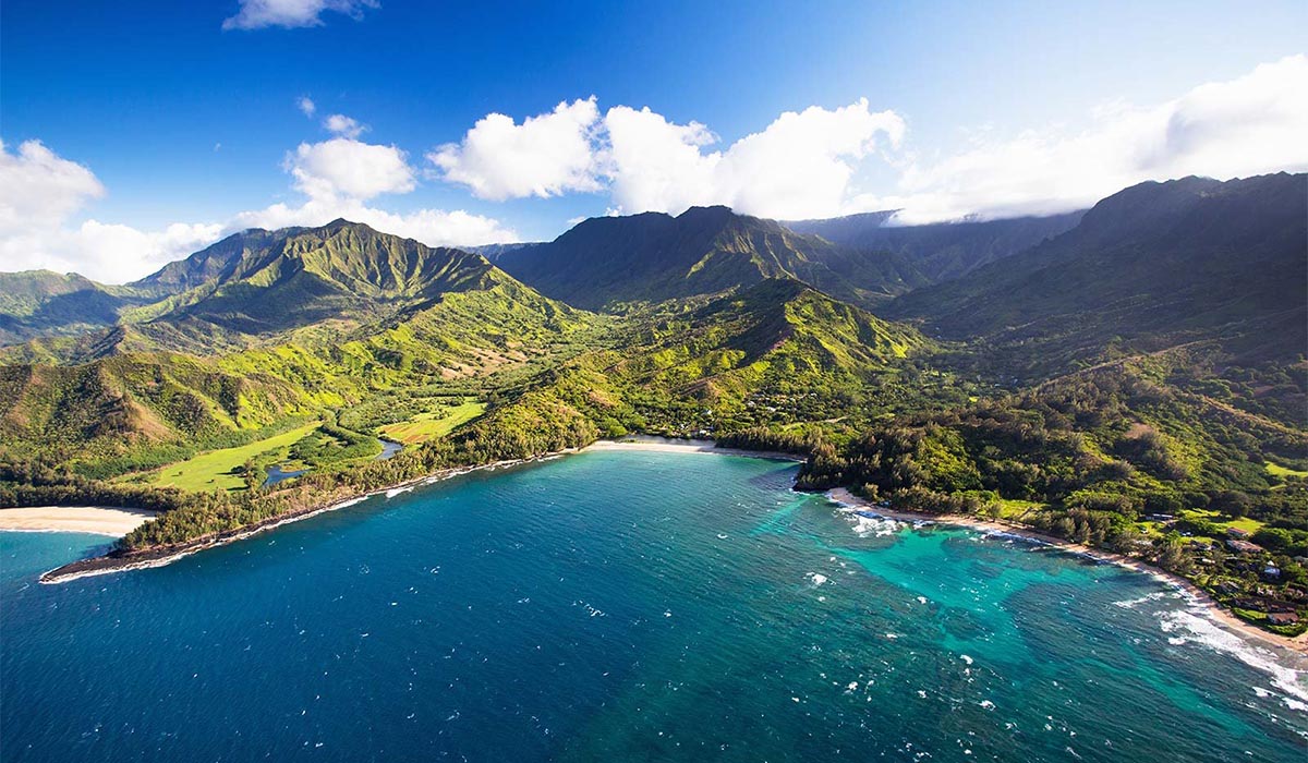 An aerial view of a lush, mountainous coastline with a crescent-shaped bay, turquoise water, and green valleys stretching inland under a bright, partly cloudy sky.
