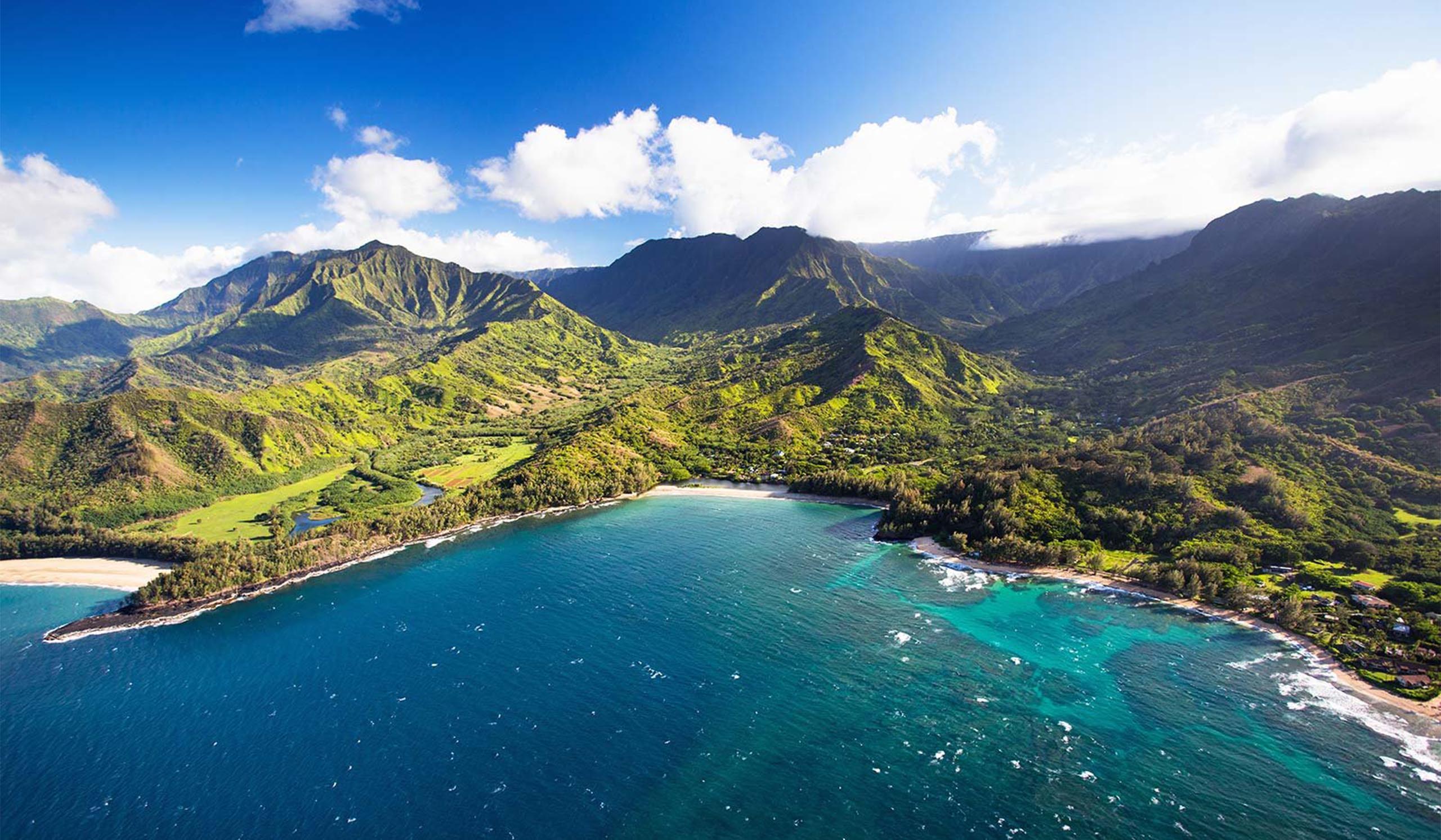An aerial view of a lush, mountainous coastline with a crescent-shaped bay, turquoise water, and green valleys stretching inland under a bright, partly cloudy sky.