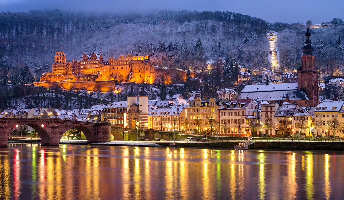 A riverside town with a lit castle on a hill glows at dusk, with reflections on the water and snow-covered rooftops.