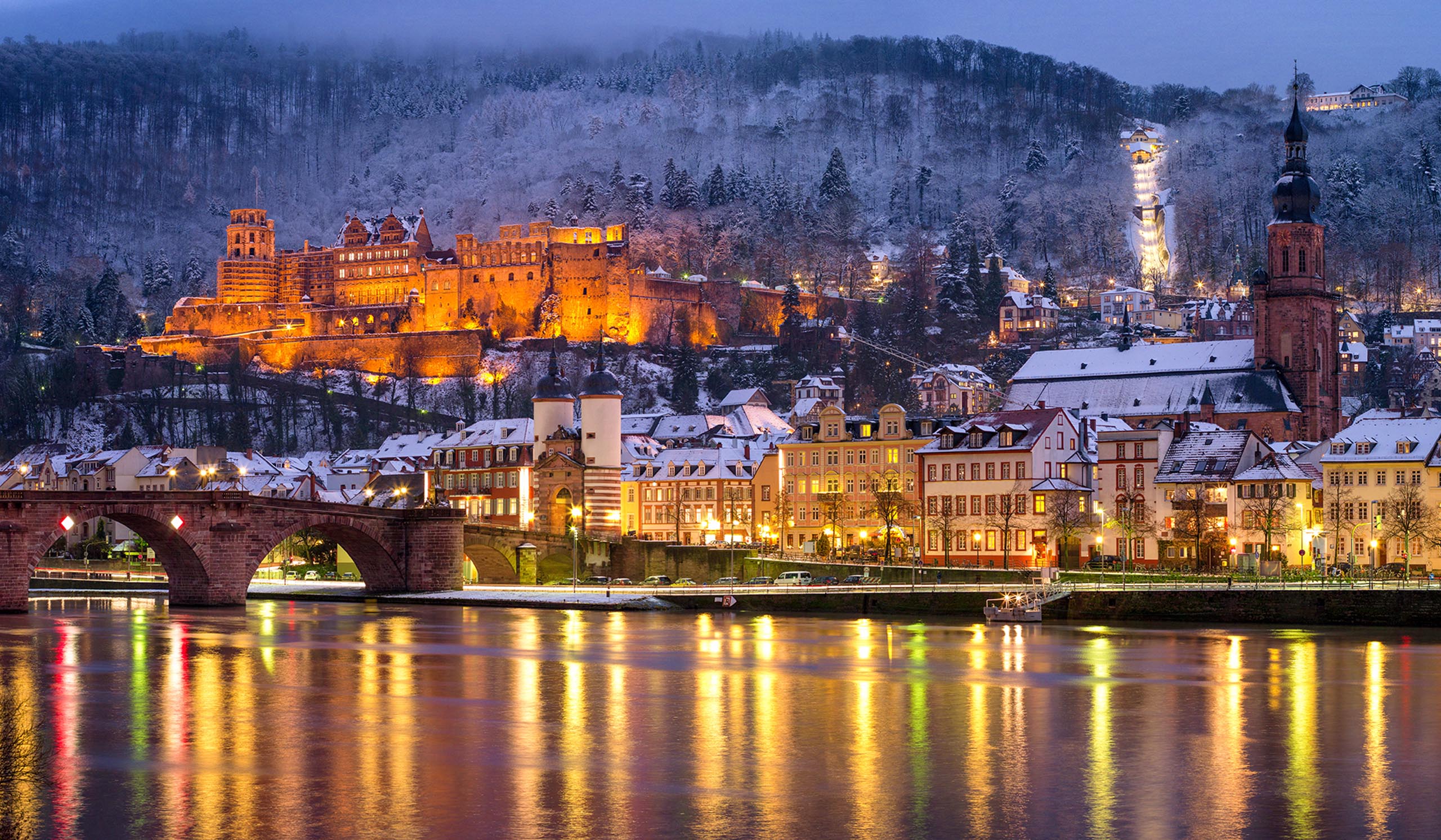 A riverside town with a lit castle on a hill glows at dusk, with reflections on the water and snow-covered rooftops.