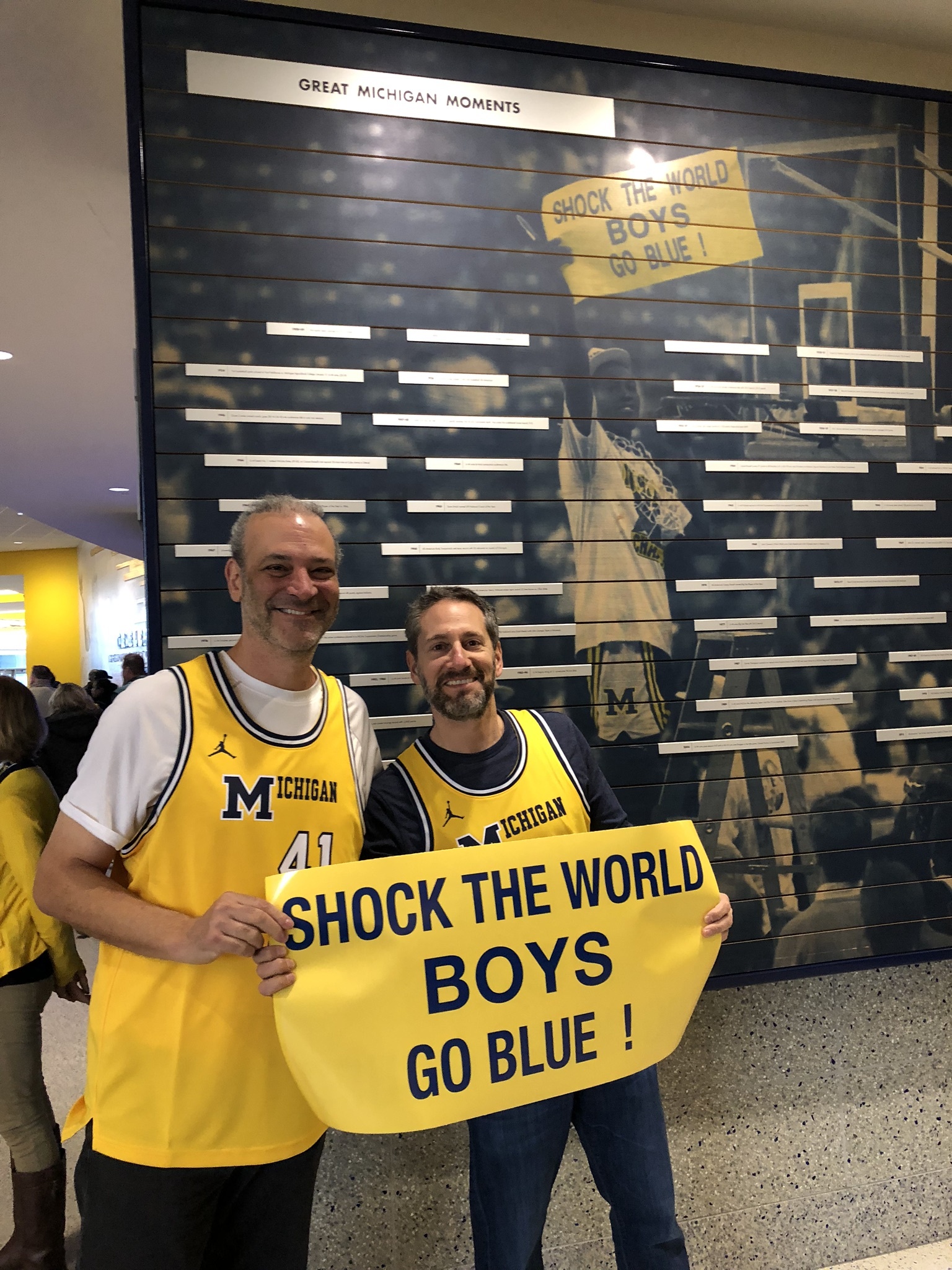 Two alums in yellow basketball jerseys pose with the "Shock the World" sign in front of the 1989 photo displayed in Crisler Center. 