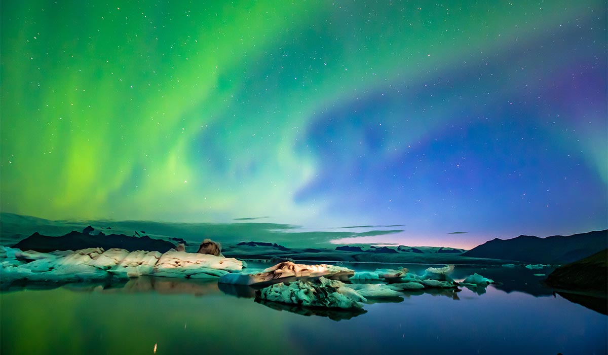 Green and blue northern lights glow over a calm lagoon with floating ice under a starry night sky.