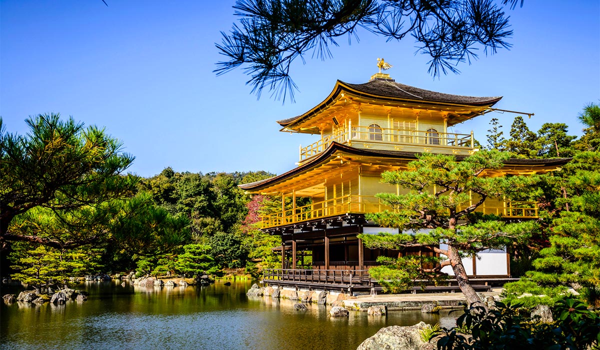 Golden temple with tiered roofs sits beside a reflective pond, surrounded by lush greenery and trees.
