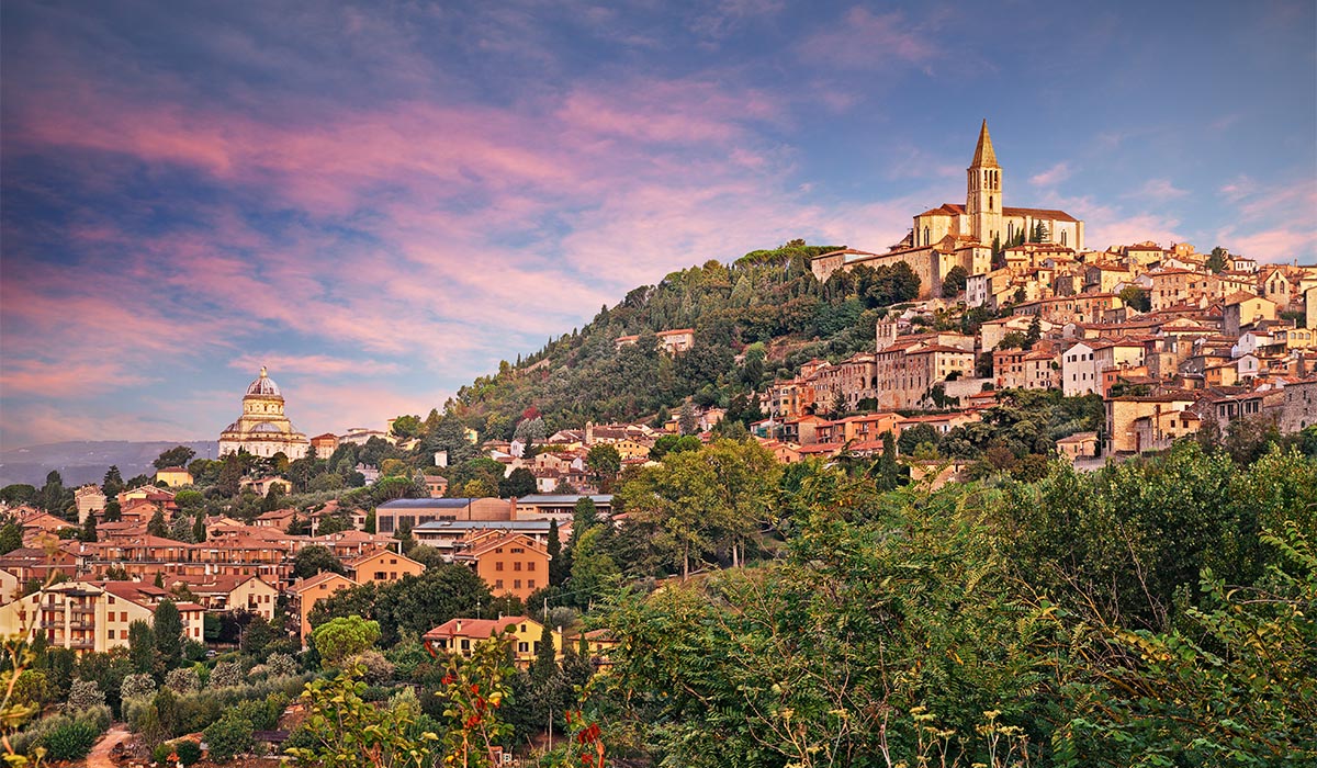 A hillside town with clustered buildings and a church sits above a green landscape under a colorful sunset sky.