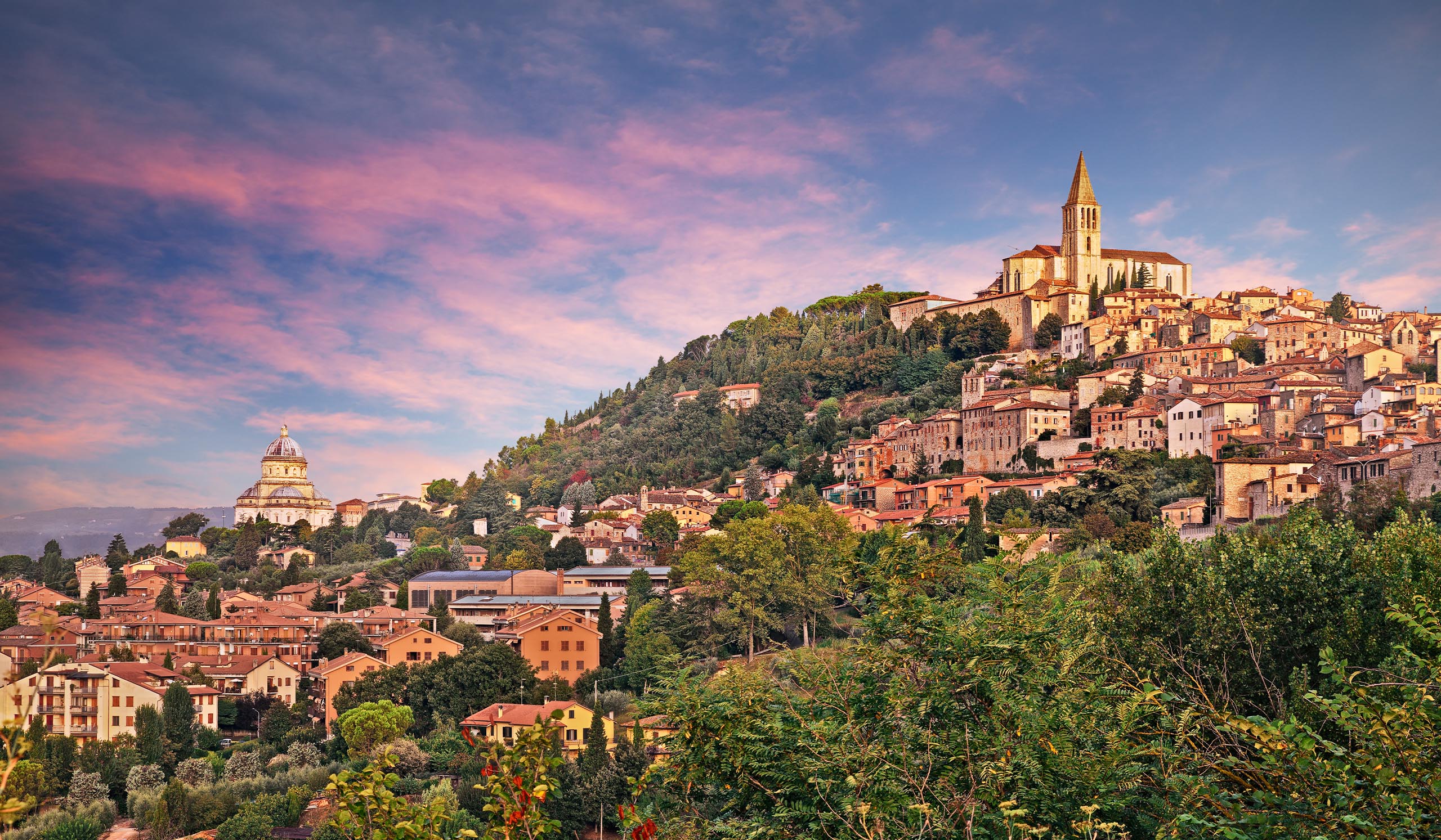 A hillside town with clustered buildings and a church sits above a green landscape under a colorful sunset sky.