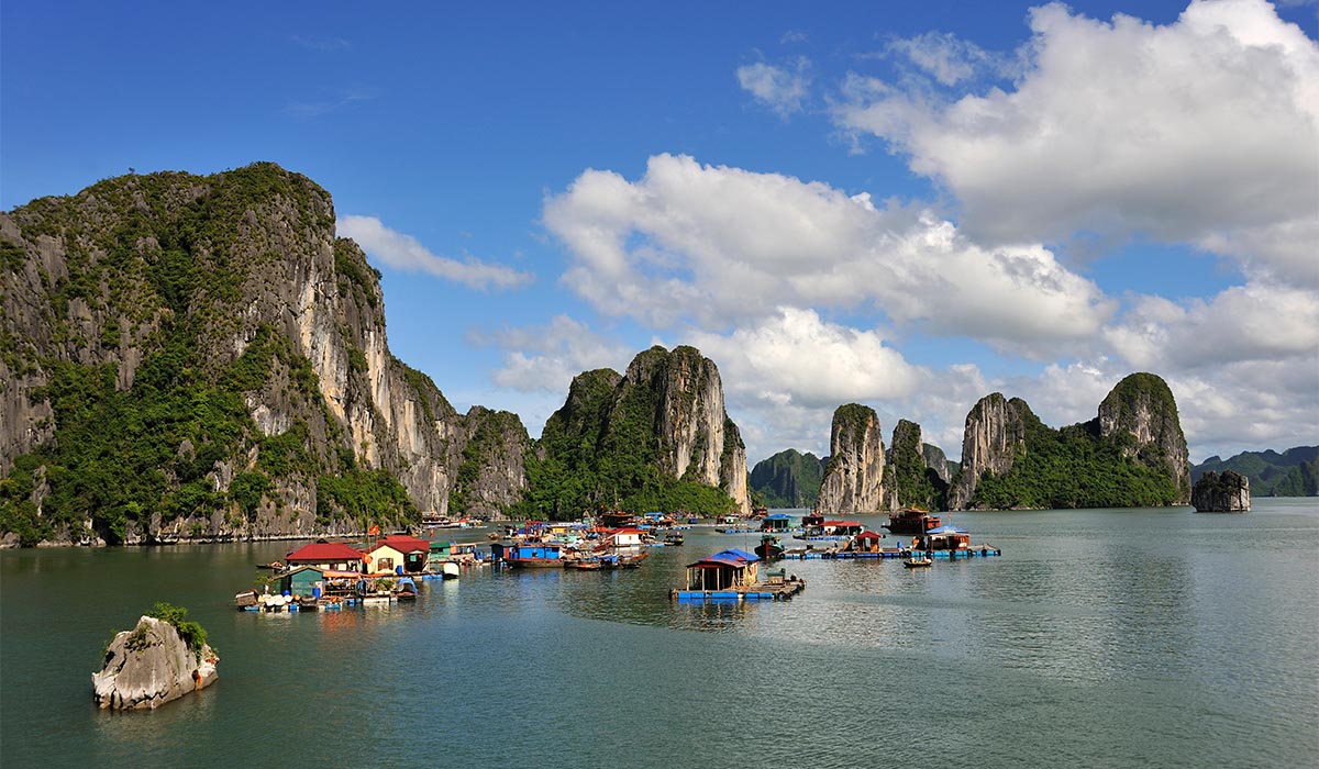 Floating village with colorful houses sits on calm water, surrounded by towering limestone cliffs under a partly cloudy sky.