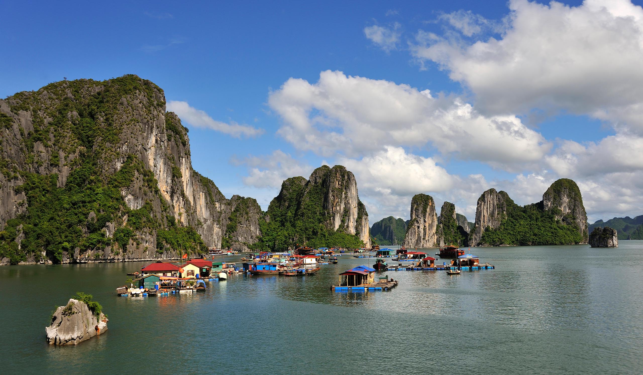 Floating village with colorful houses sits on calm water, surrounded by towering limestone cliffs under a partly cloudy sky.
