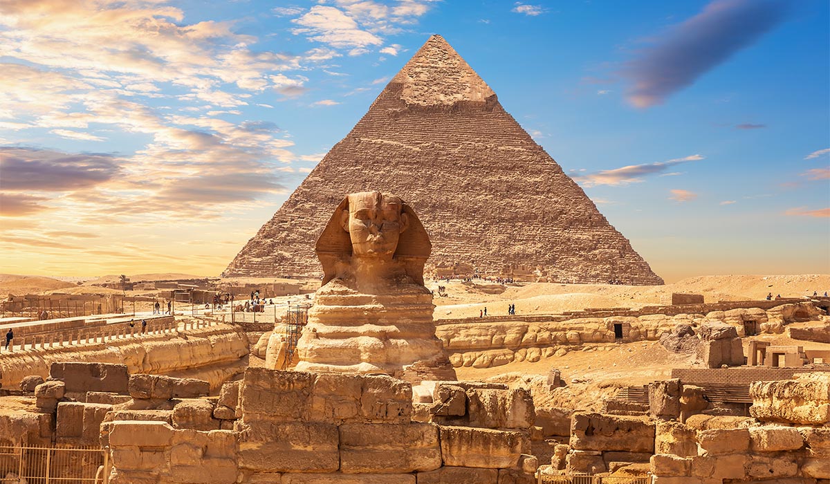 The Great Sphinx stands in front of a large pyramid in the desert under a partly cloudy sky.