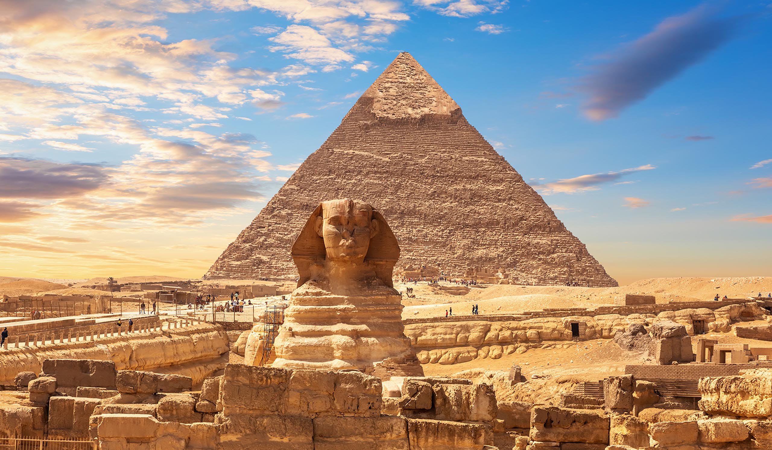 The Great Sphinx stands in front of a large pyramid in the desert under a partly cloudy sky.