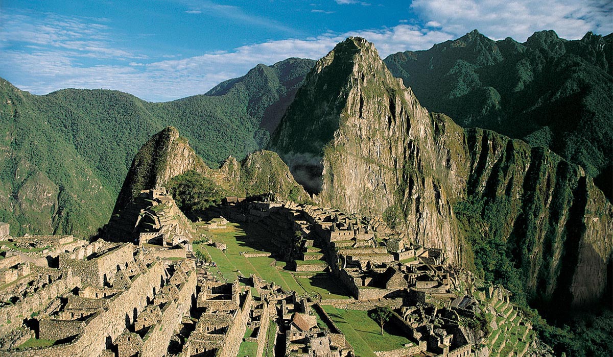 Ancient stone ruins of Machu Picchu sit among steep, green mountains under a bright blue sky.
