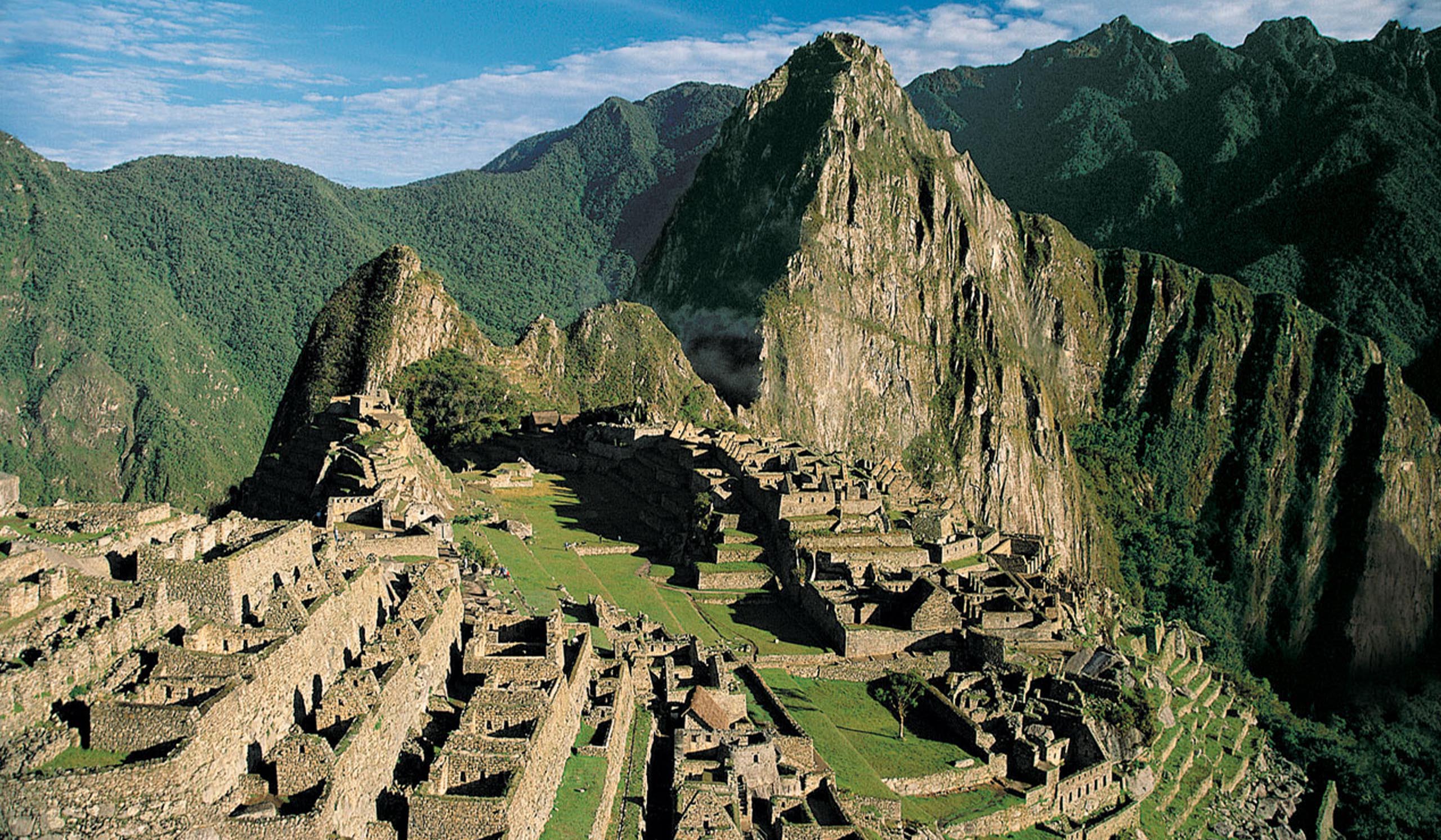 Ancient stone ruins of Machu Picchu sit among steep, green mountains under a bright blue sky.