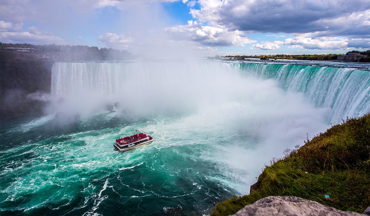 A powerful view of Niagara Falls with a tour boat full of visitors in red ponchos approaching the base, surrounded by mist and turquoise, churning water under a partly cloudy sky.