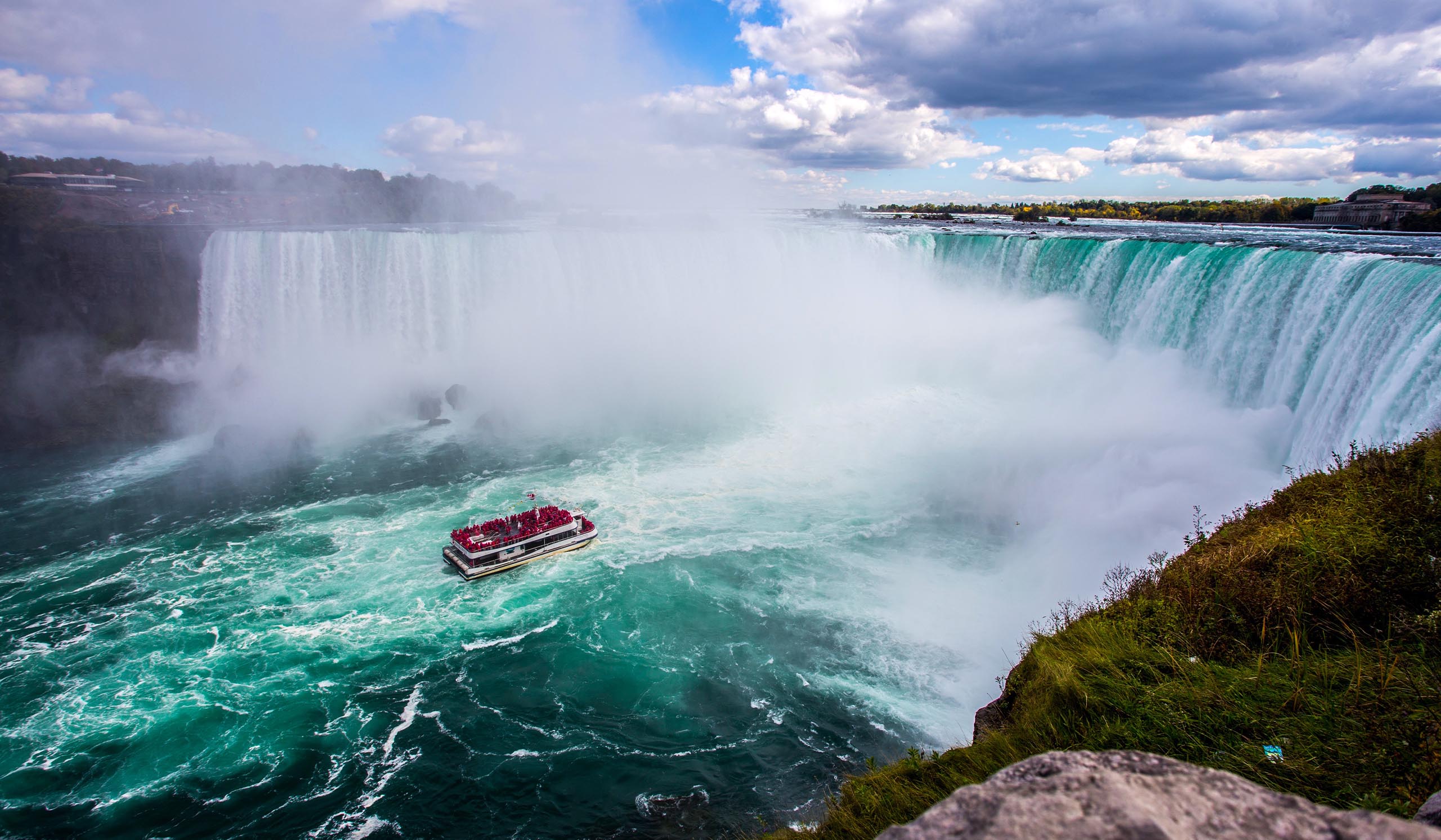 A powerful view of Niagara Falls with a tour boat full of visitors in red ponchos approaching the base, surrounded by mist and turquoise, churning water under a partly cloudy sky.