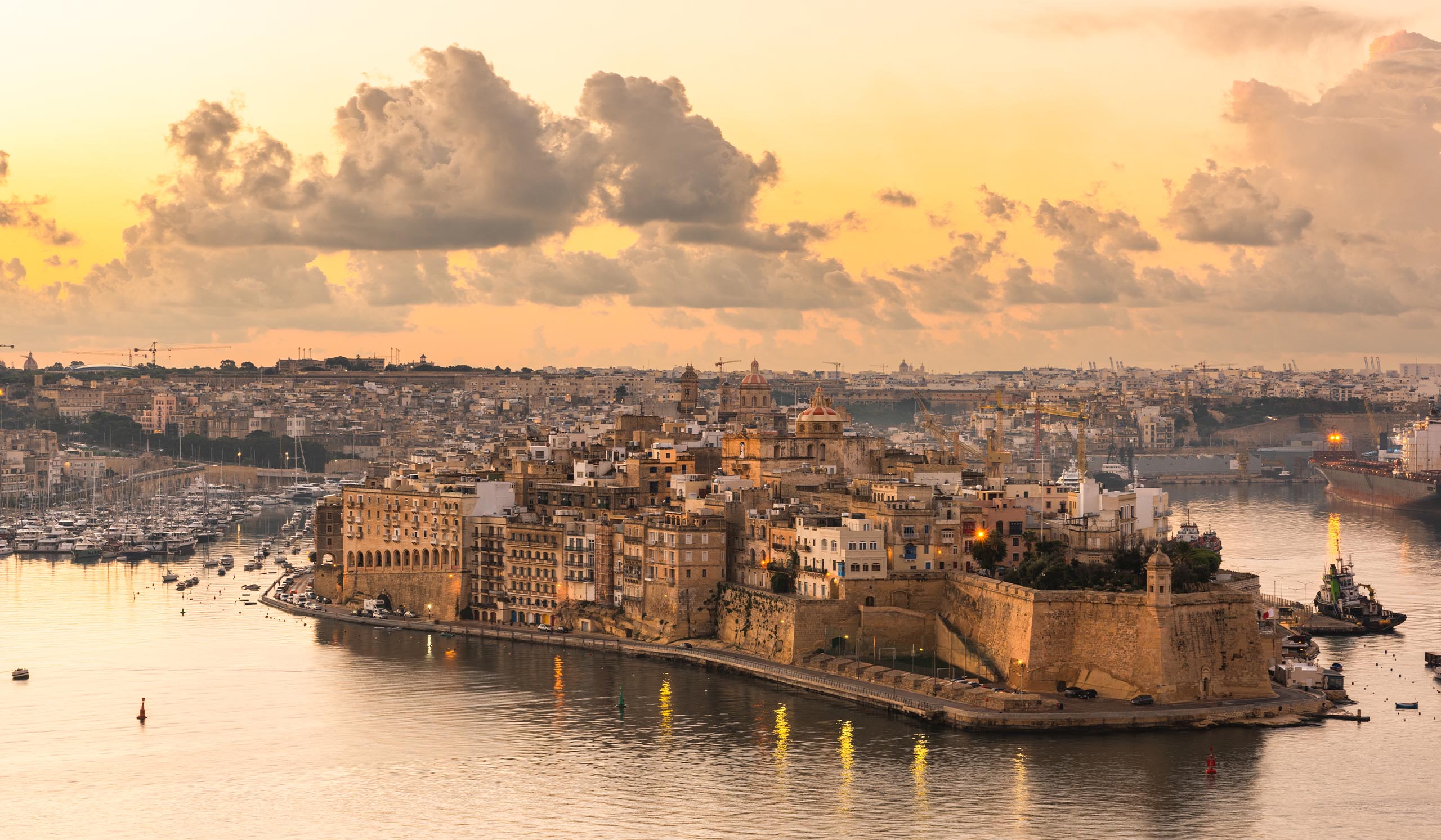 A historic coastal city with stone buildings and fort walls sits along a harbor at sunset, with boats in the water and a golden sky overhead.