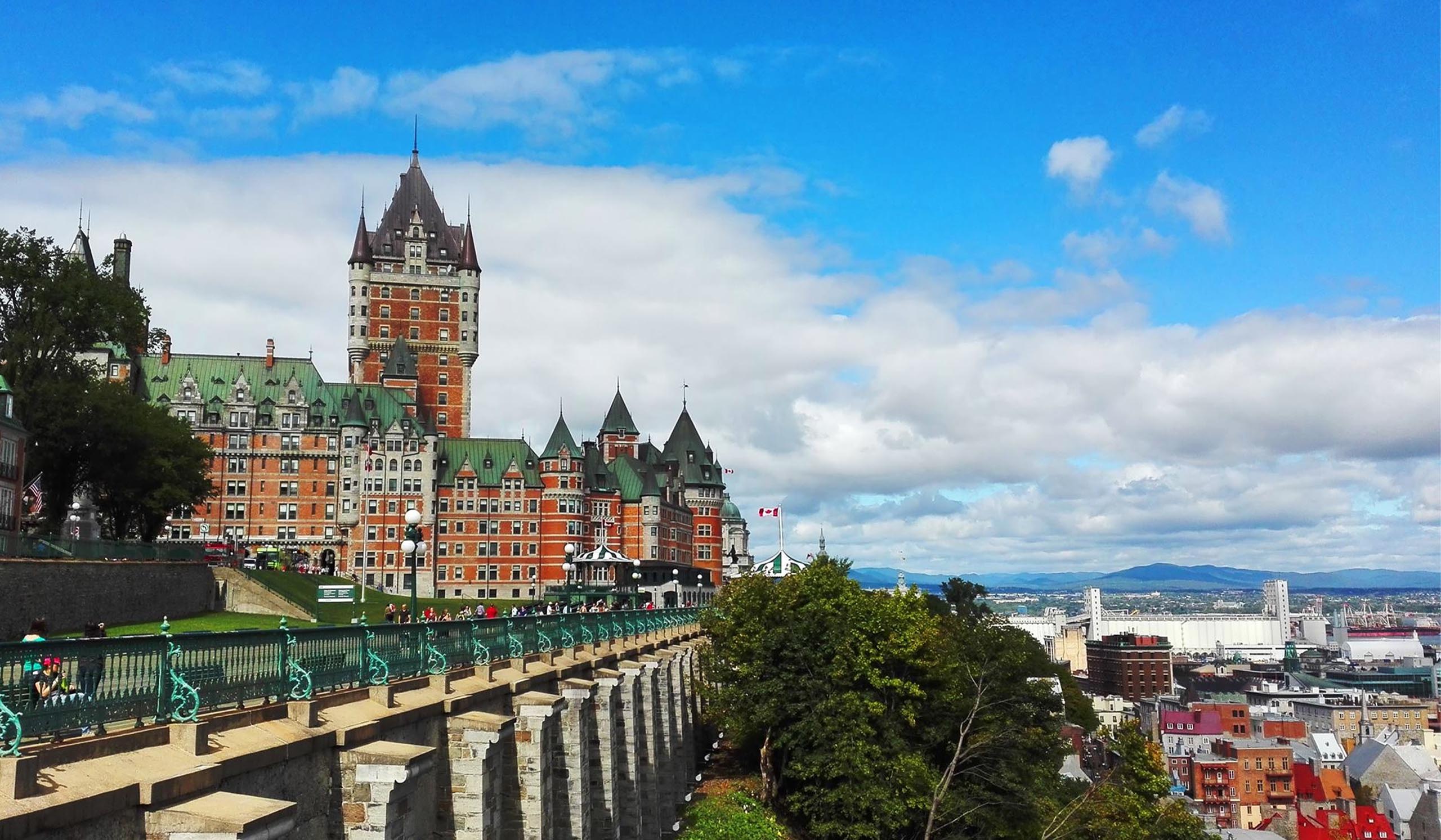 A grand, castle-like historic hotel with green roofs and a tall central tower overlooks a riverside city from a scenic bluff under a partly cloudy sky.