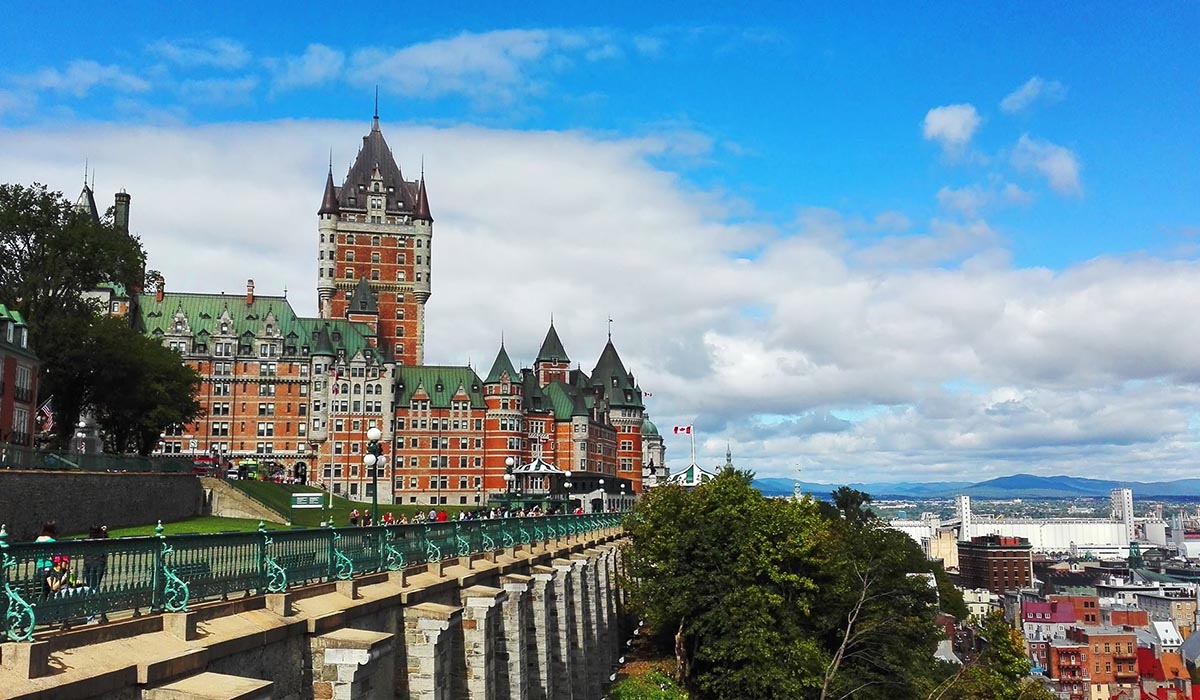 A grand, castle-like historic hotel with green roofs and a tall central tower overlooks a riverside city from a scenic bluff under a partly cloudy sky.