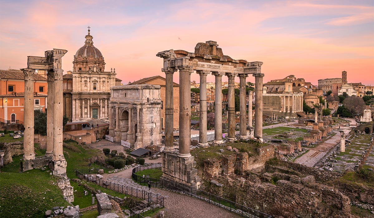 Ancient Roman ruins with tall columns stand in a historic site at sunset, with buildings in the background.
