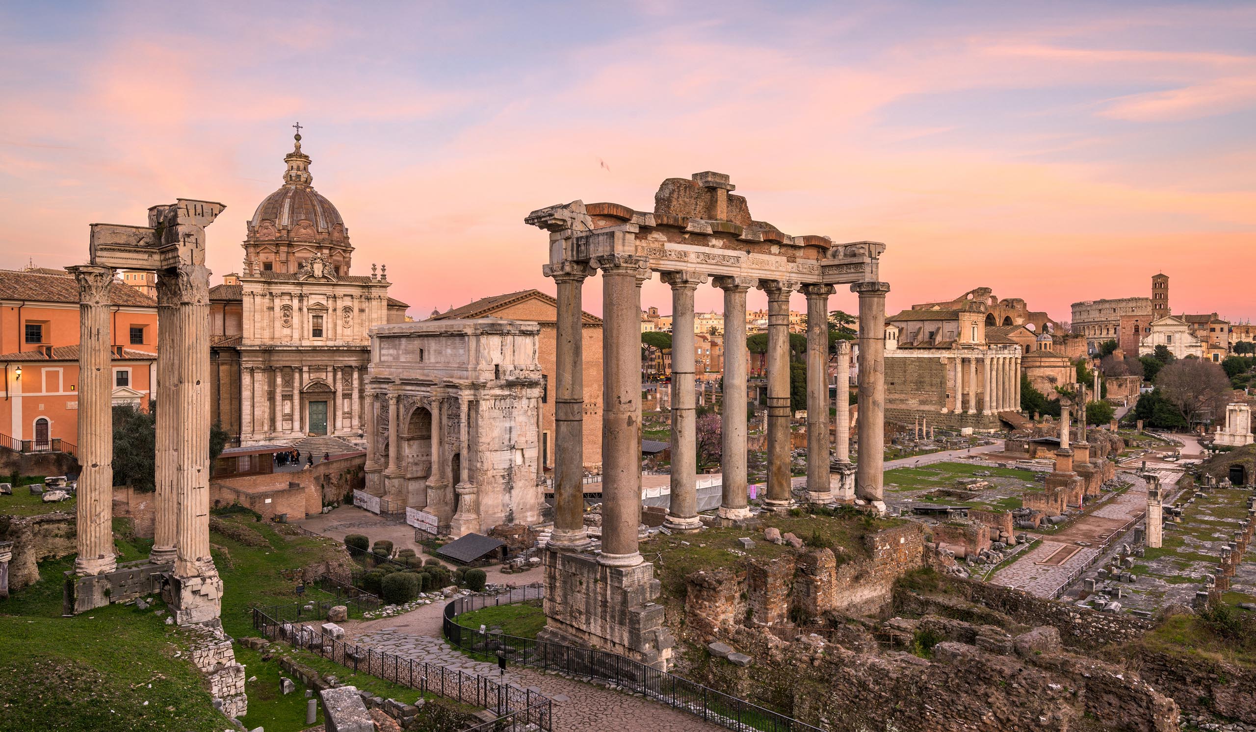 Ancient Roman ruins with tall columns stand in a historic site at sunset, with buildings in the background.