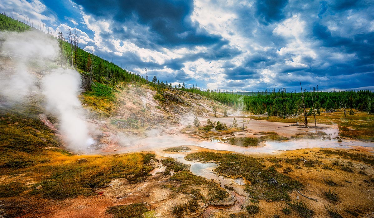 Steaming geothermal landscape with vents and hot springs spread across a colorful, mineral-stained basin, surrounded by forested hills under a dramatic, cloud-filled sky.