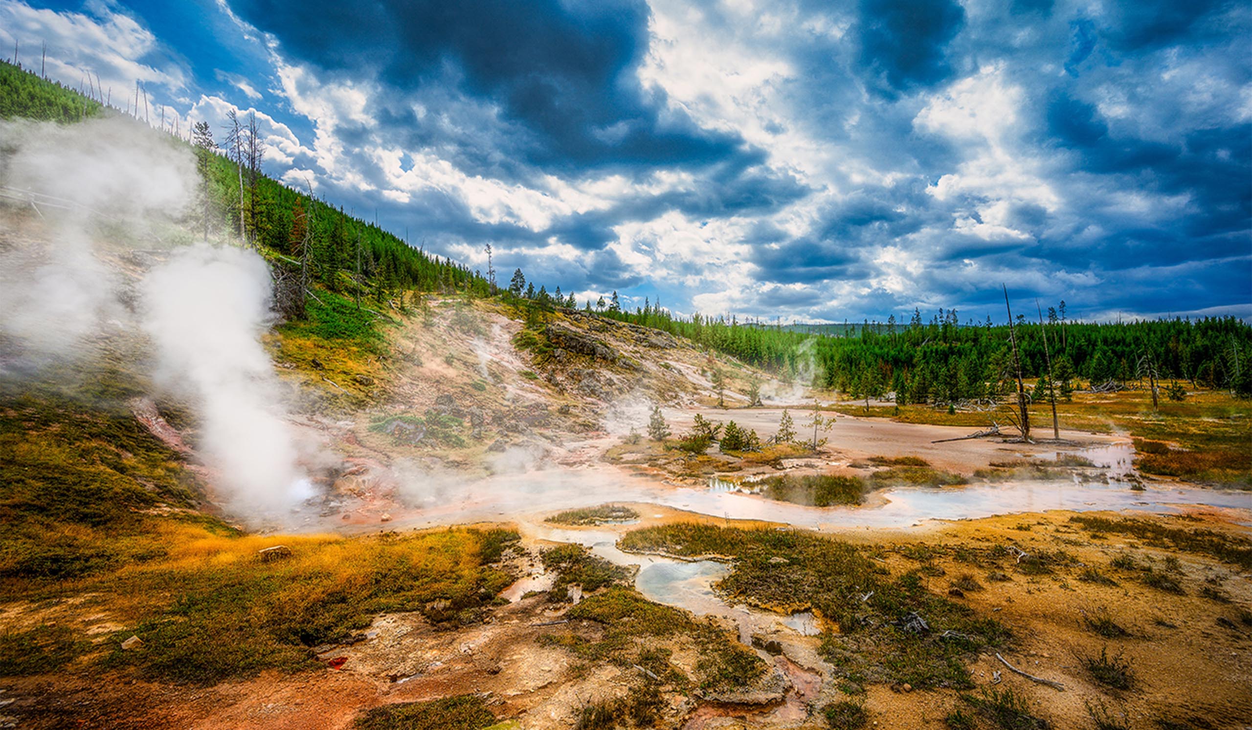 Steaming geothermal landscape with vents and hot springs spread across a colorful, mineral-stained basin, surrounded by forested hills under a dramatic, cloud-filled sky.