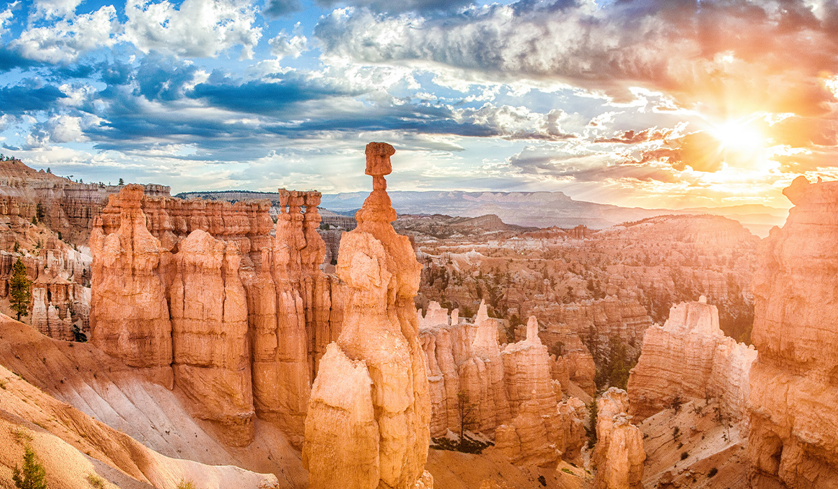 Sunlight streams over towering orange rock formations in a desert canyon, with dramatic clouds filling the sky.