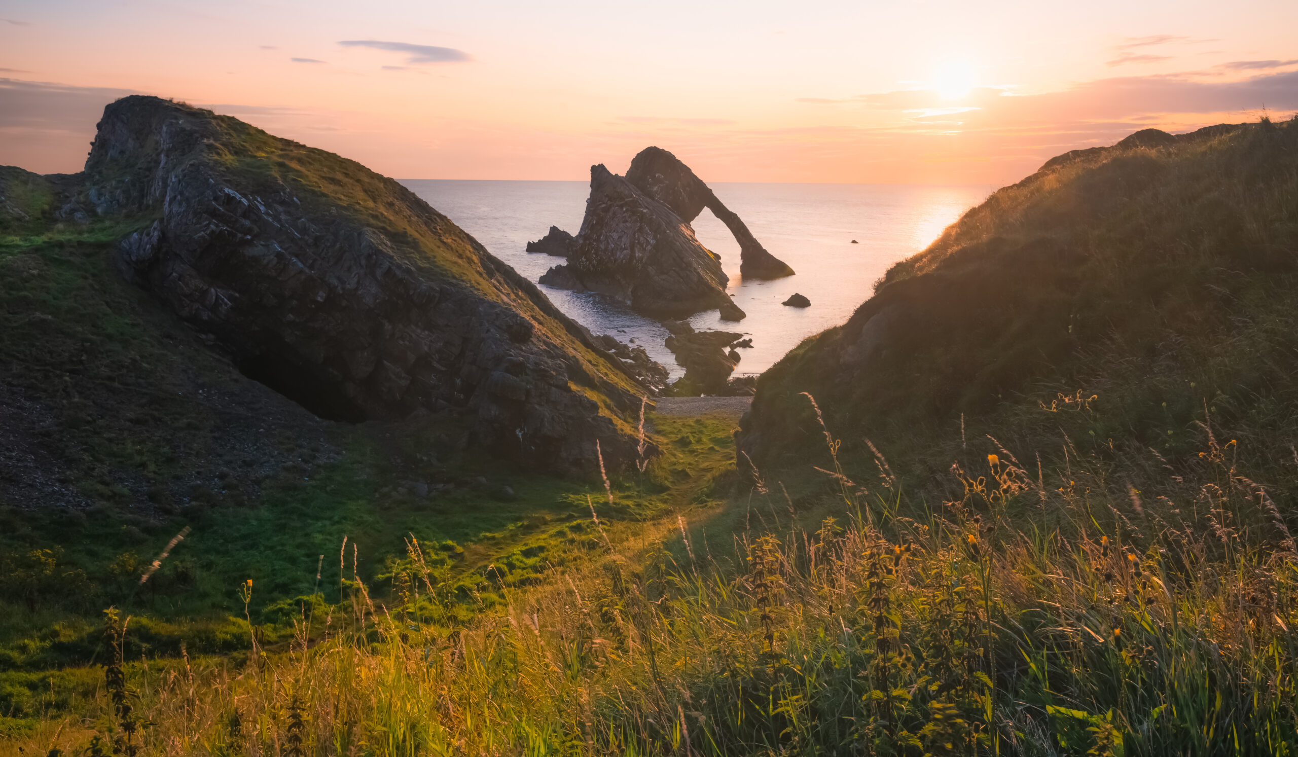 Dramatic coastal landscape at sunset with rugged cliffs and a natural rock arch rising from the sea, framed by grassy hills glowing in warm, golden light.