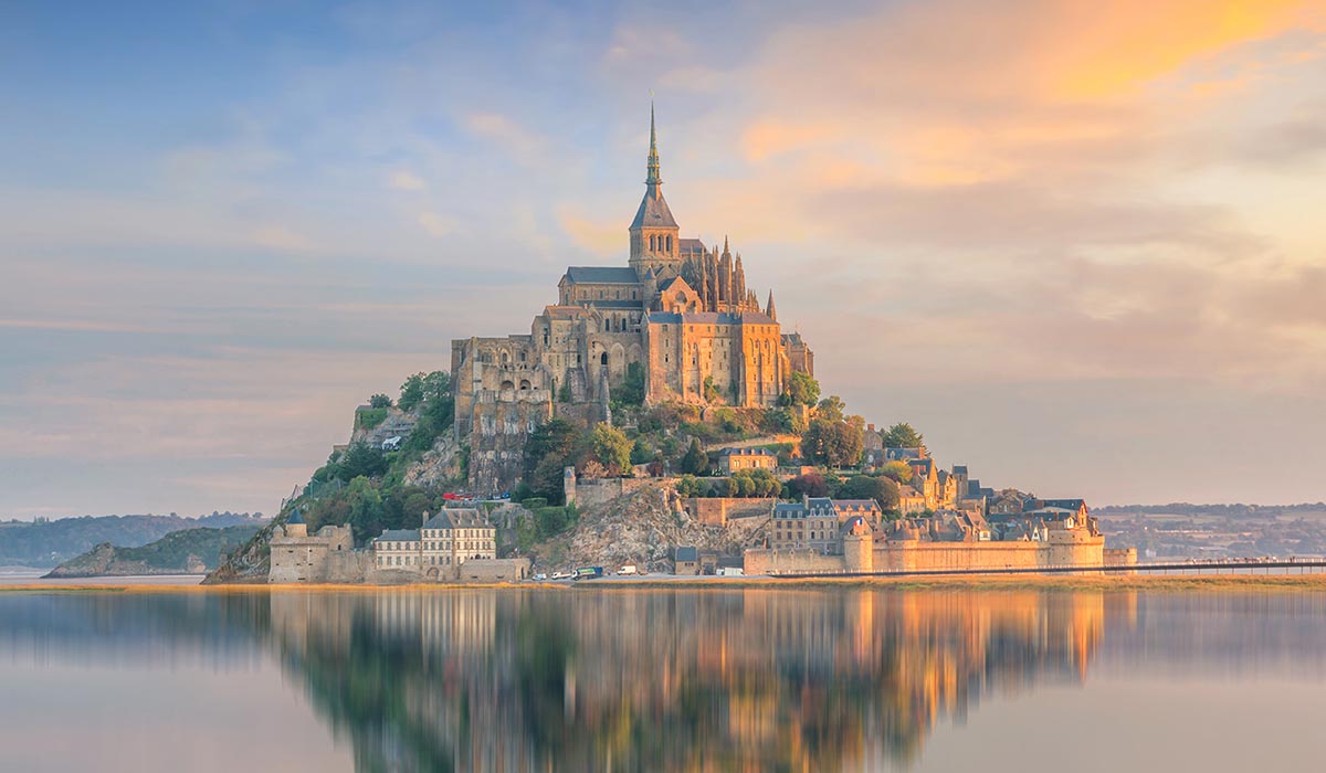 Historic Mont-Saint-Michel sits on a rocky island, reflected in calm water at sunset.