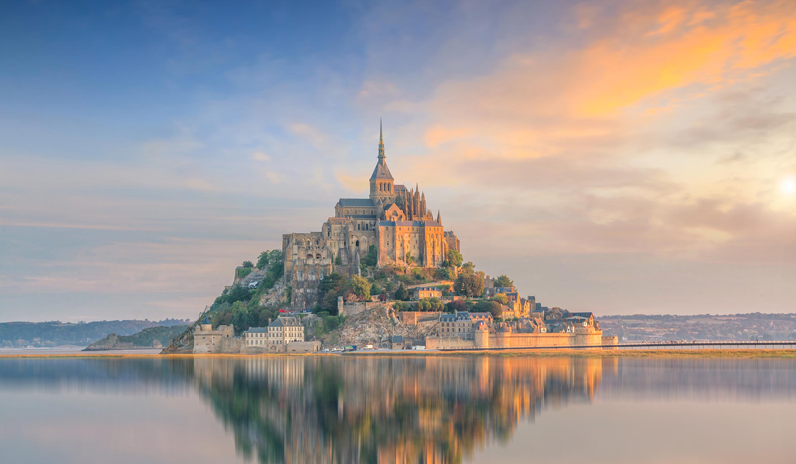 Historic Mont-Saint-Michel sits on a rocky island, reflected in calm water at sunset.