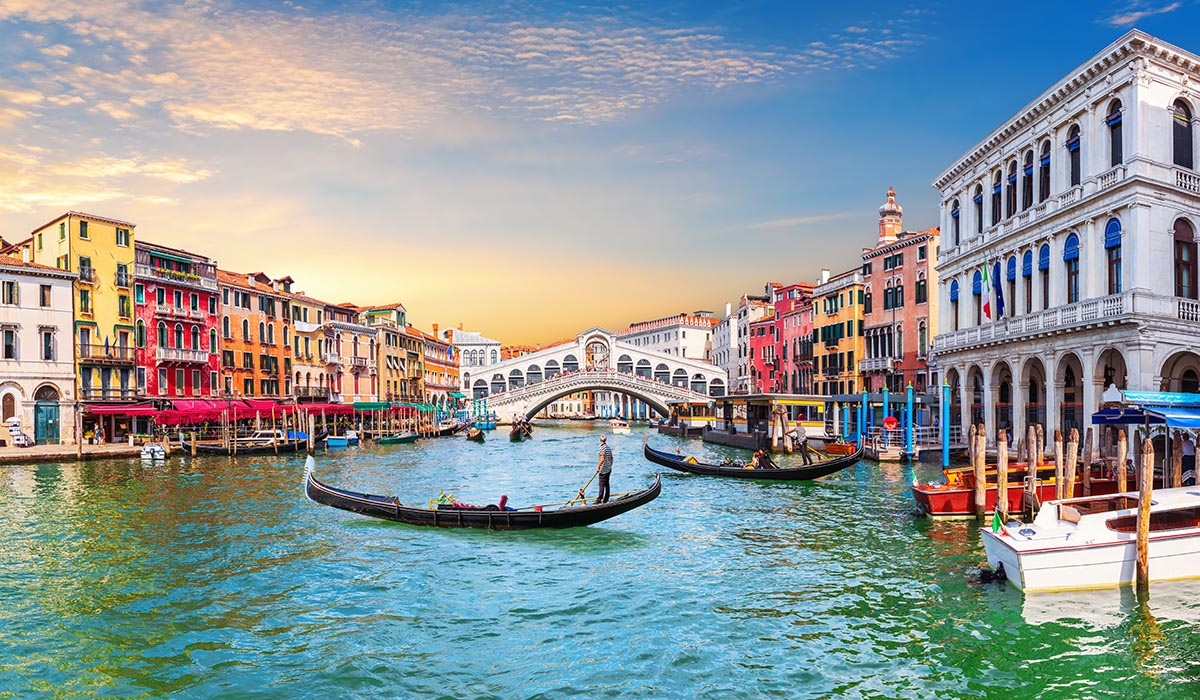 Gondolas glide along a canal lined with colorful buildings, with a bridge in the background under a bright sky.