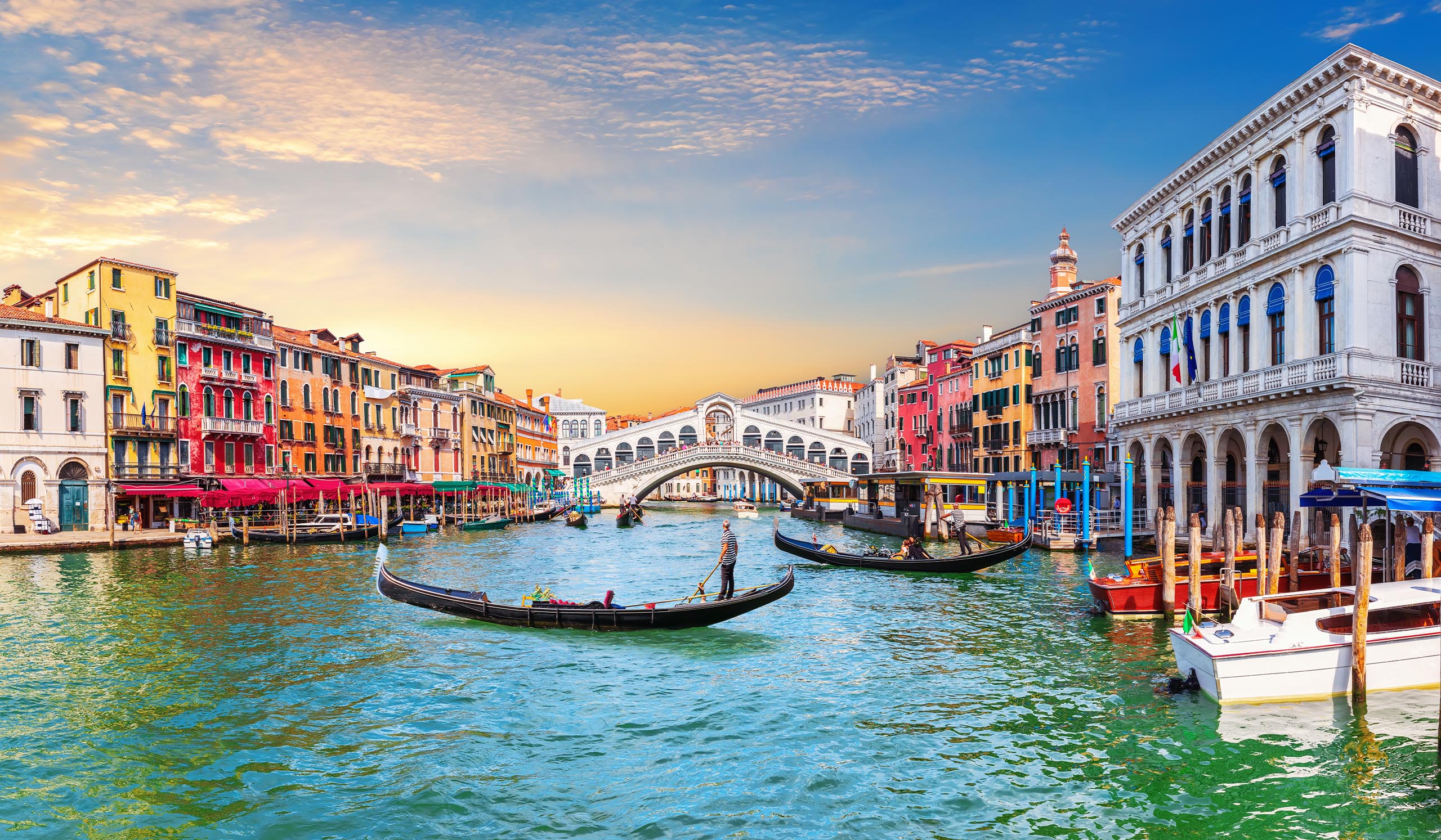 Gondolas glide along a canal lined with colorful buildings, with a bridge in the background under a bright sky.