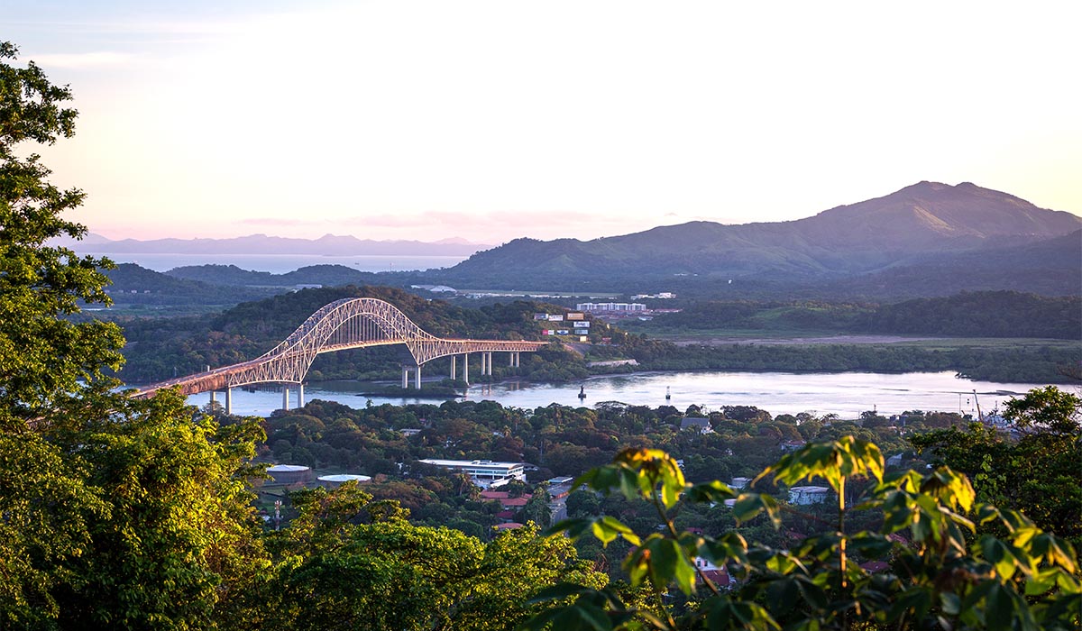 A large arch bridge spans a river through a green landscape, with hills and water in the distance.