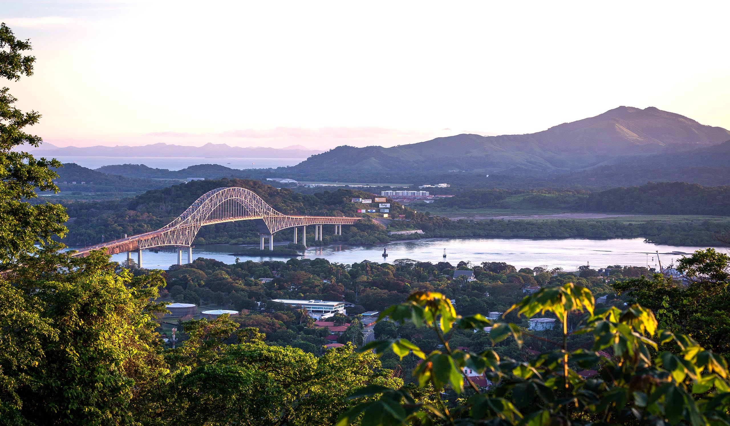 A large arch bridge spans a river through a green landscape, with hills and water in the distance.