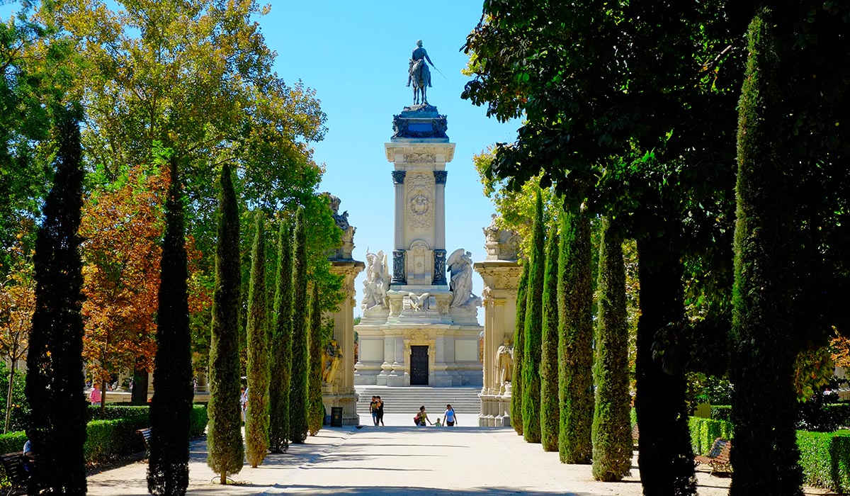 Tree-lined walkway leads to a tall monument topped with a statue, with a few people gathered at its base under a clear blue sky.